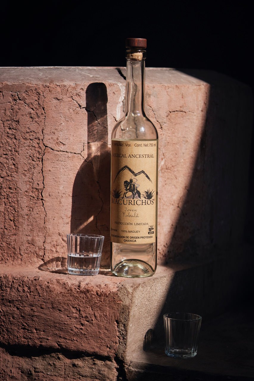 A glass bottle of mezcal, labeled 'Mezcal Ancestral Macurichos Joven Tobalá' with a cork stopper, positioned on a rough pink stone surface, with a small glass of clear liquid beside it. The background is dark, with shadows cast on the stone.