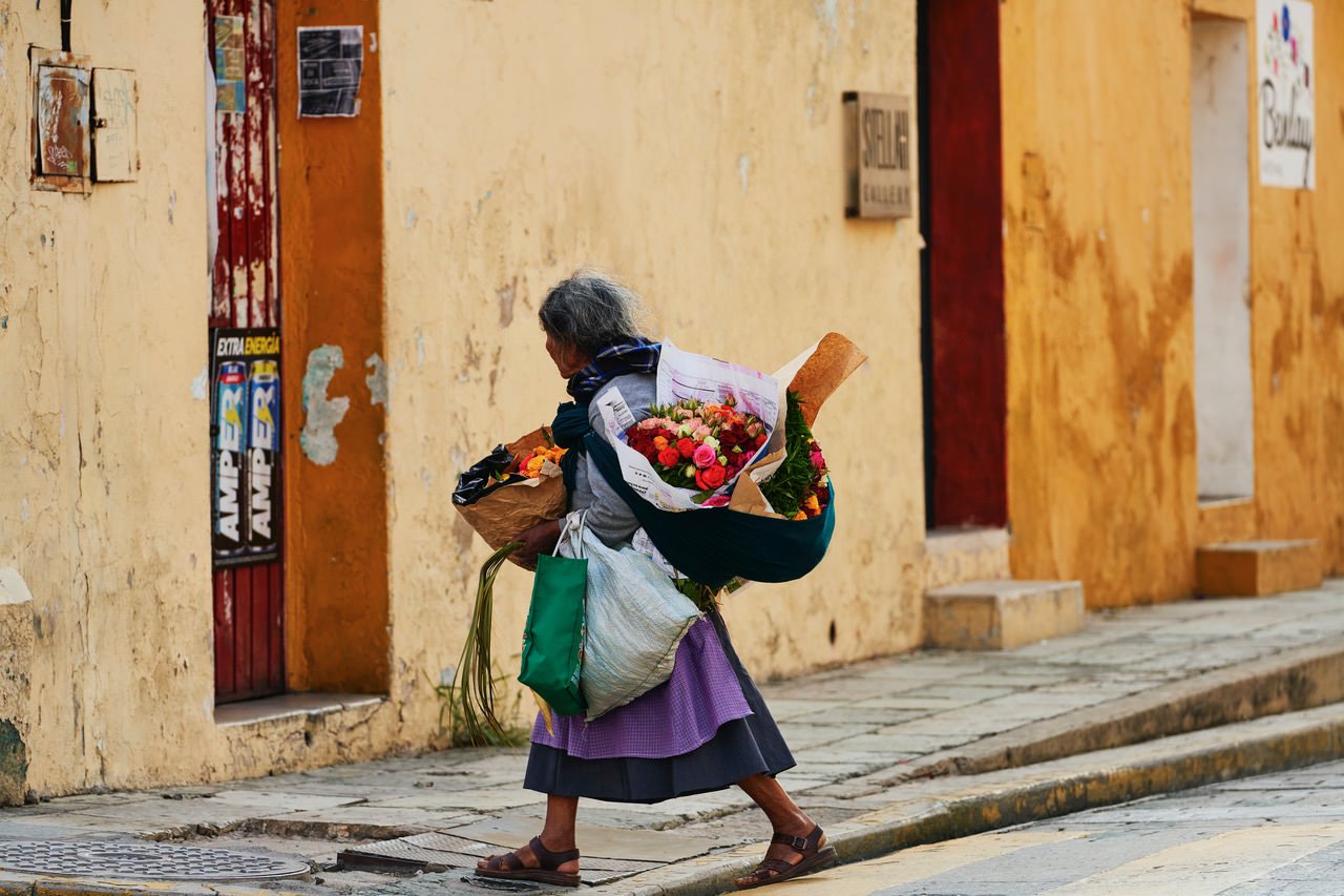 An elderly woman carrying bouquets of colorful flowers and large bags walking along a cobblestone street outside a weathered yellow wall.