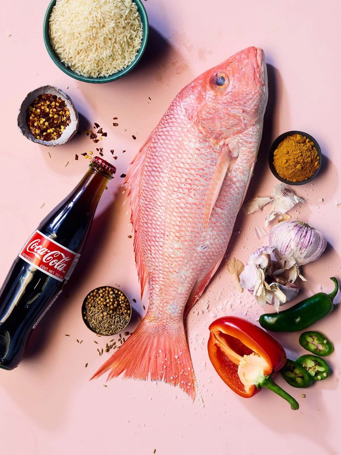 Fresh fish surrounded by rice, chili flakes, curry powder, garlic, jalapeño peppers, tomato, and a Coca-Cola bottle, on a pink surface.