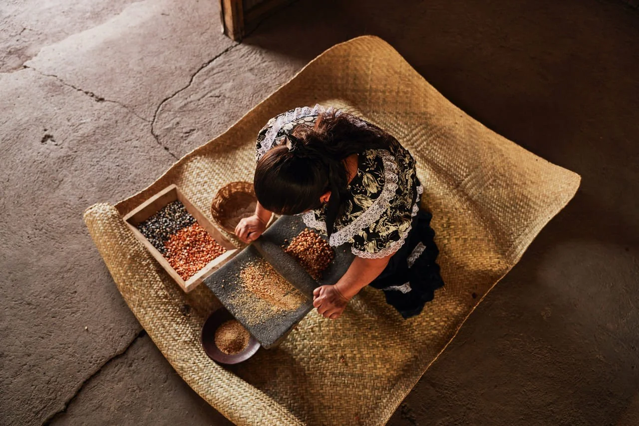 A woman sitting on a woven mat, sorting beans on a black tray and in a wooden box.