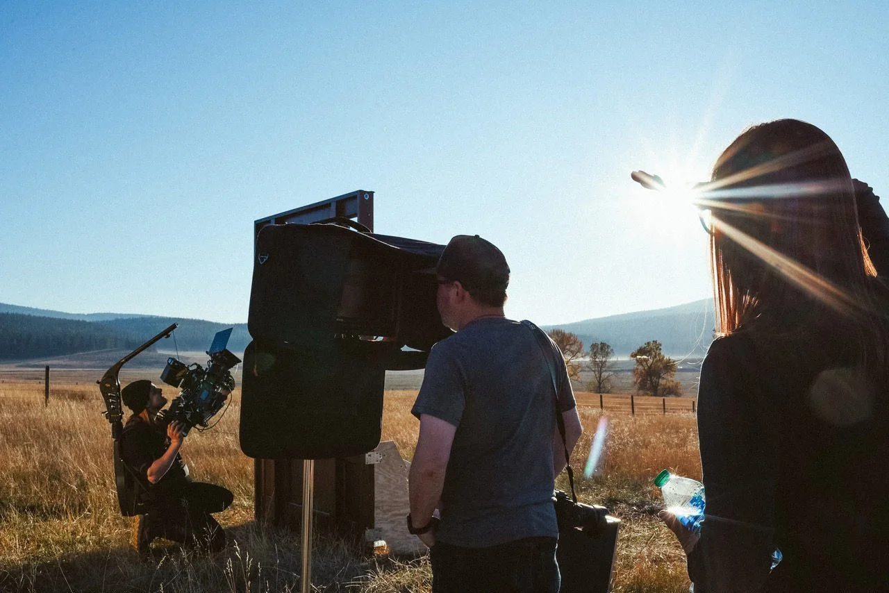 Filmmakers working outdoors in a field with mountains in the background, using a camera mounted on a tripod and large equipment, during sunset.