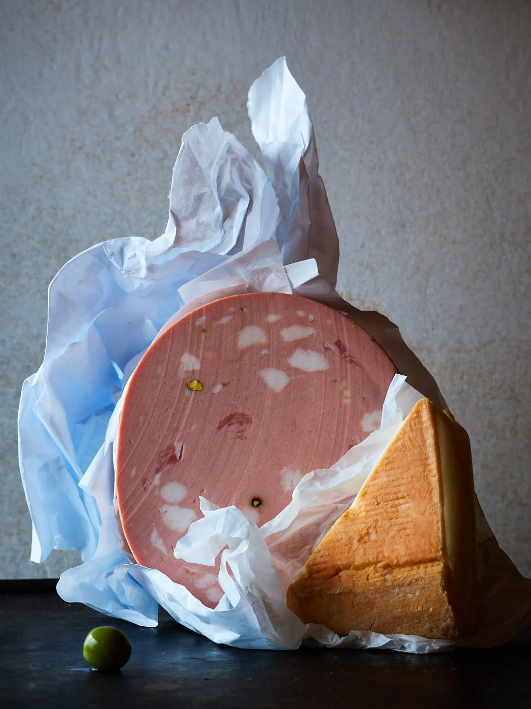 A block of sliced ham with a wedge of cheese on a dark surface, with a small green grape nearby, all against a neutral background.