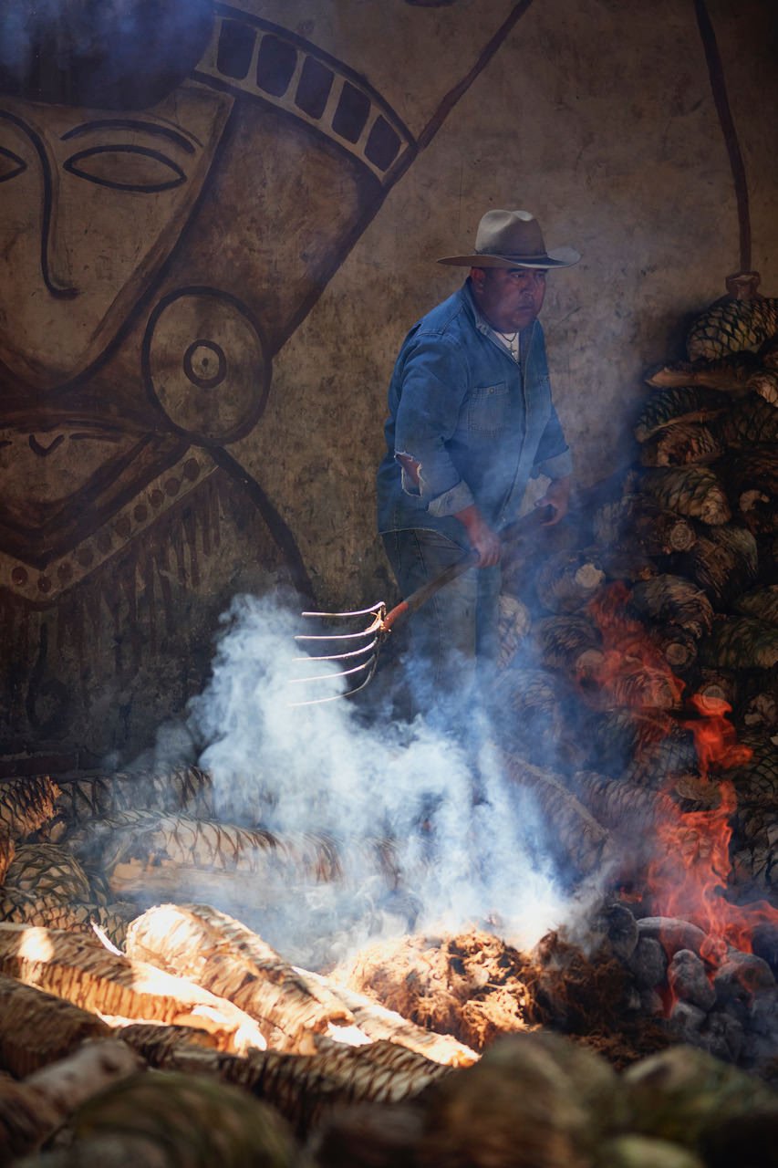 A man wearing a hat and denim shirt standing over a fire with burning logs, smoke rising, and a large mural of a face and geometric designs on the wall behind him.