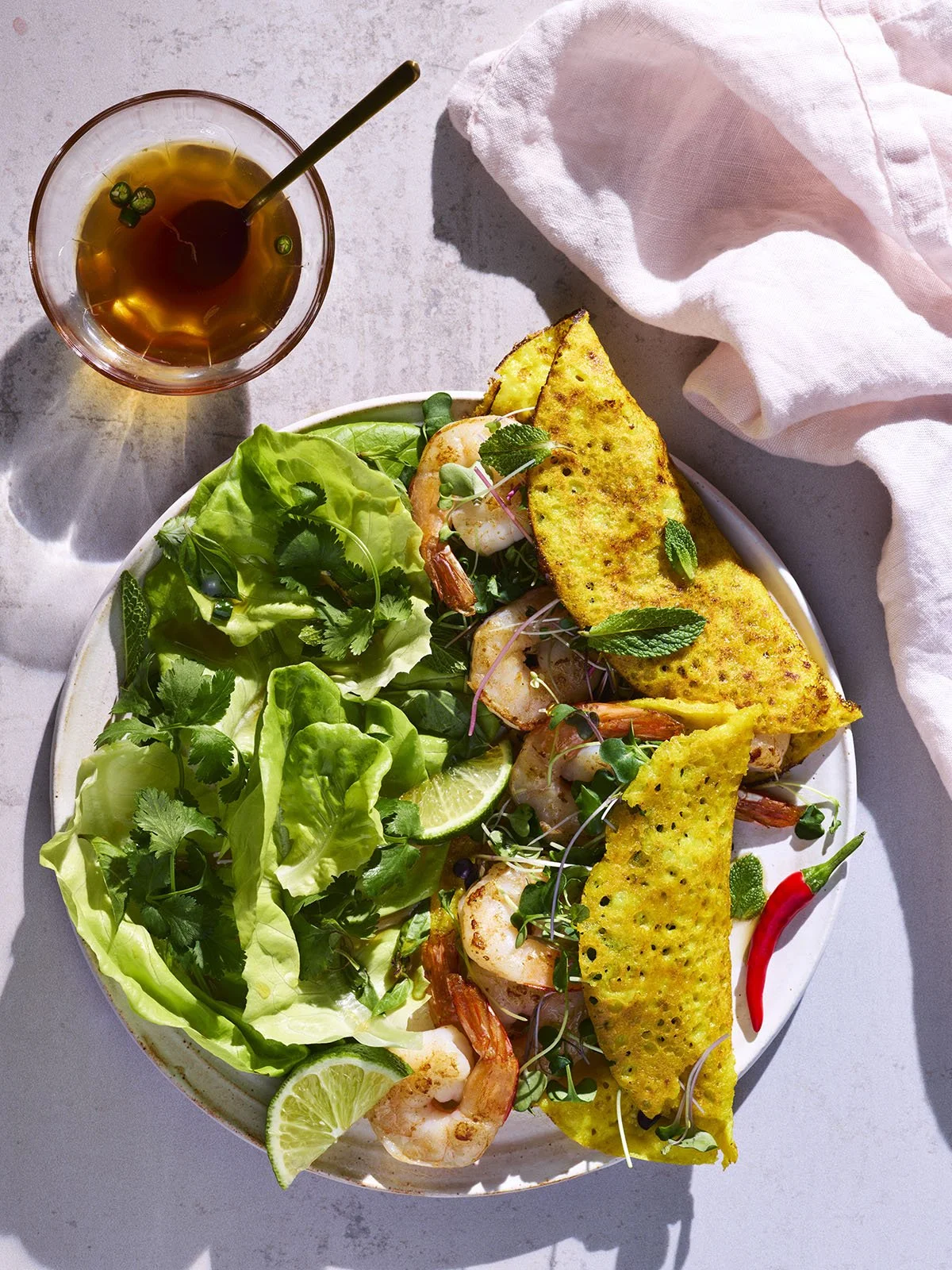 A plate of shrimp and green vegetables with lime slices, topped with edible flowers, and garnished with herbs, alongside two pieces of toasted yellow bread or flatbread, and a glass of iced tea with a straw on a light-colored surface with a pink clot