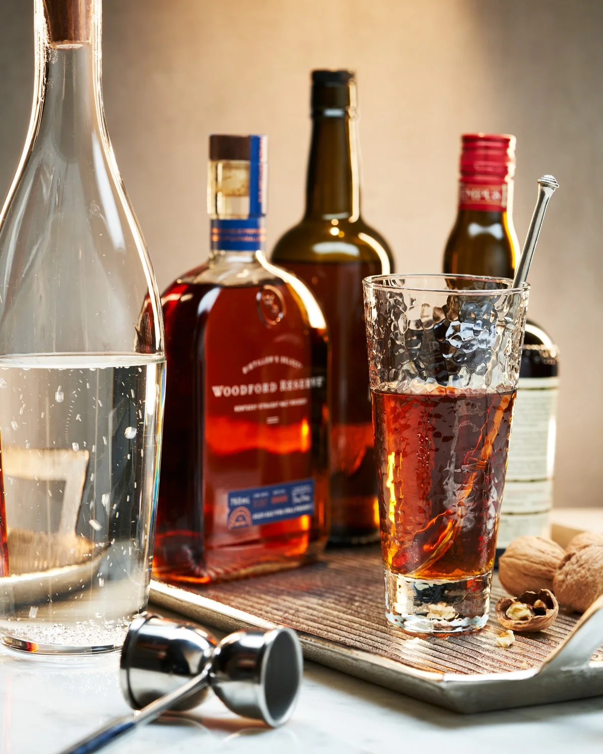 A glass of whiskey and a glass of water on a table with bottles of alcohol and walnuts in the background.
