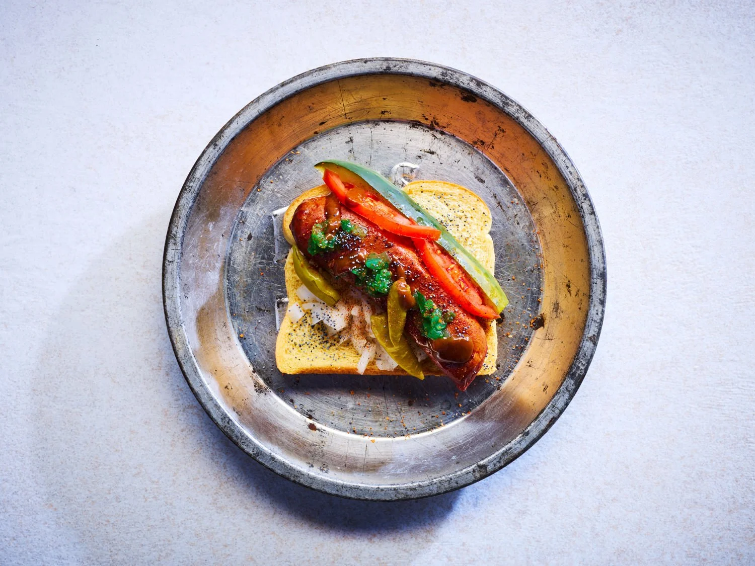 A hot dog with toppings such as tomato, pickles, chopped onions, and green relish on a toasted bun, served on a metallic tray against a light background.