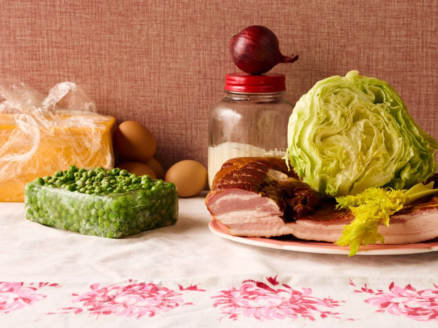 A still life of fresh groceries including green peas, eggs, a block of cheese, a head of lettuce, bacon, and a bottle of milk on a table with a pink textured wall in the background.