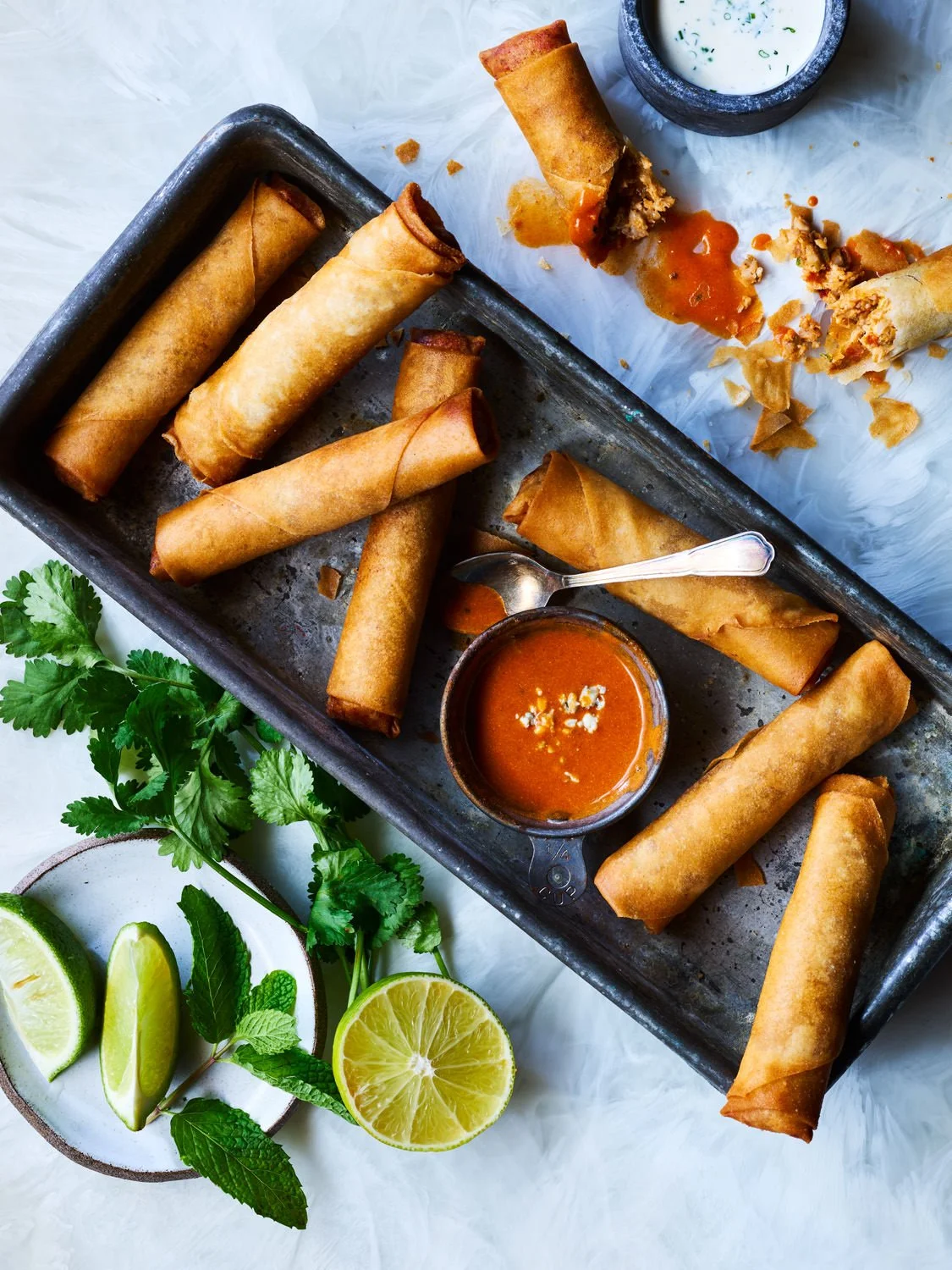 A tray of uncooked spring rolls with a small bowl of dipping sauce, scattered on crumpled white parchment paper, with lime wedges, fresh cilantro, and mint leaves nearby.