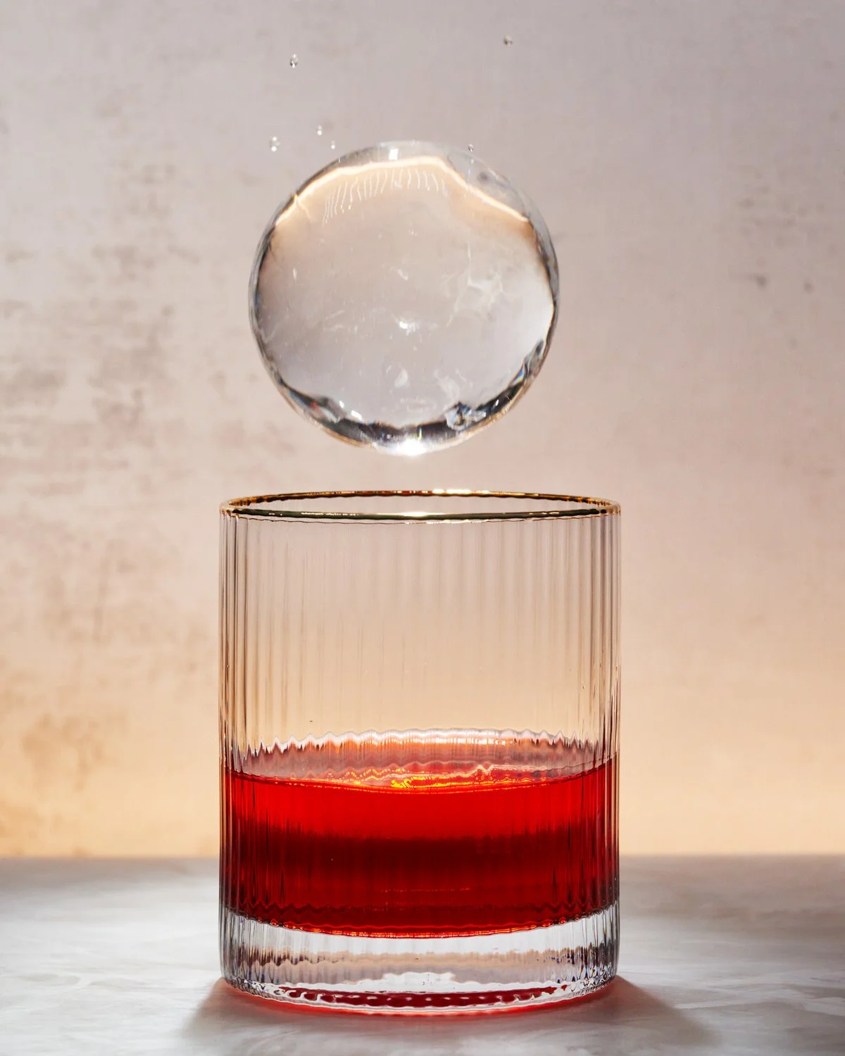 A clear glass tumbler with red liquid inside. A large ice cube is falling toward the glass, captured mid-air. The background is neutral and out of focus.