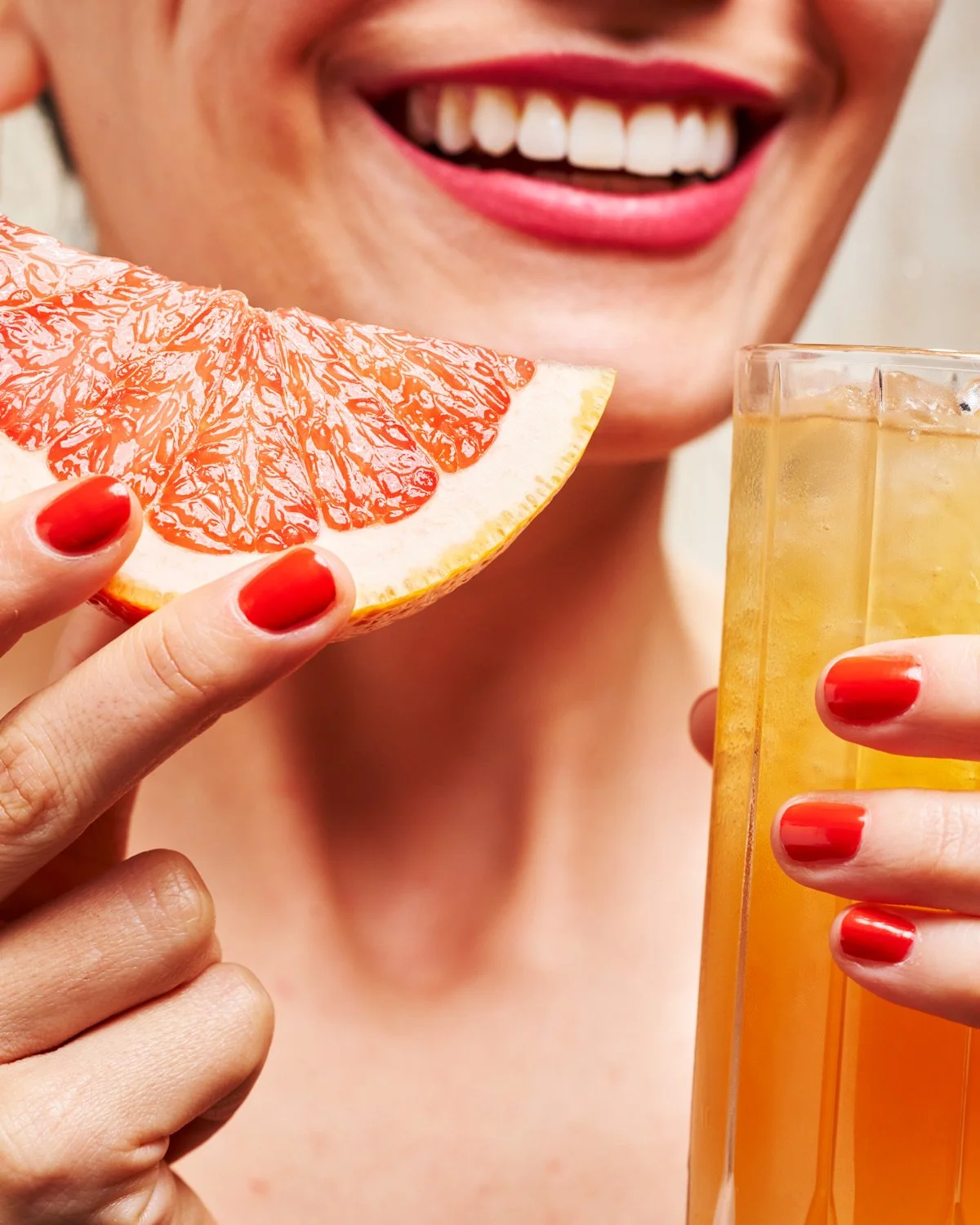 A woman with red lipstick and red nail polish holds a slice of grapefruit near her mouth and a tall glass of orange-colored beverage with ice.