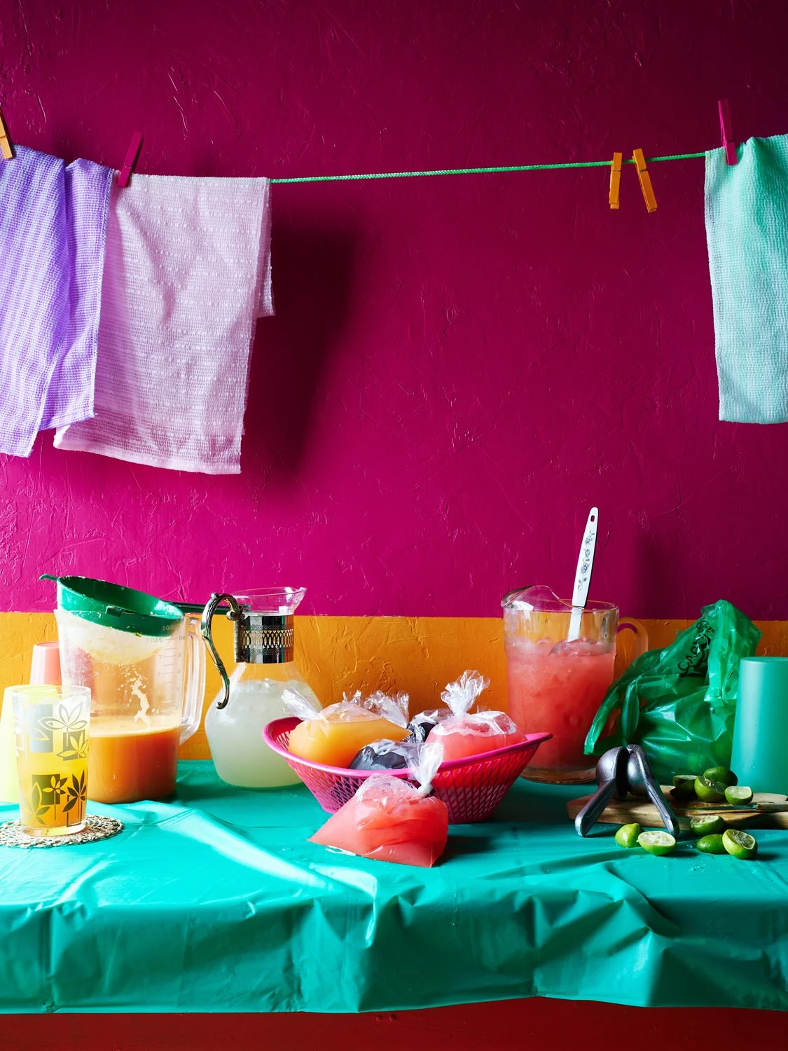 Colorful kitchen or dining area with pink and yellow wall, green tablecloth, and washing towels hanging on a line; various drinks, soap, and a green plastic bag on the table.