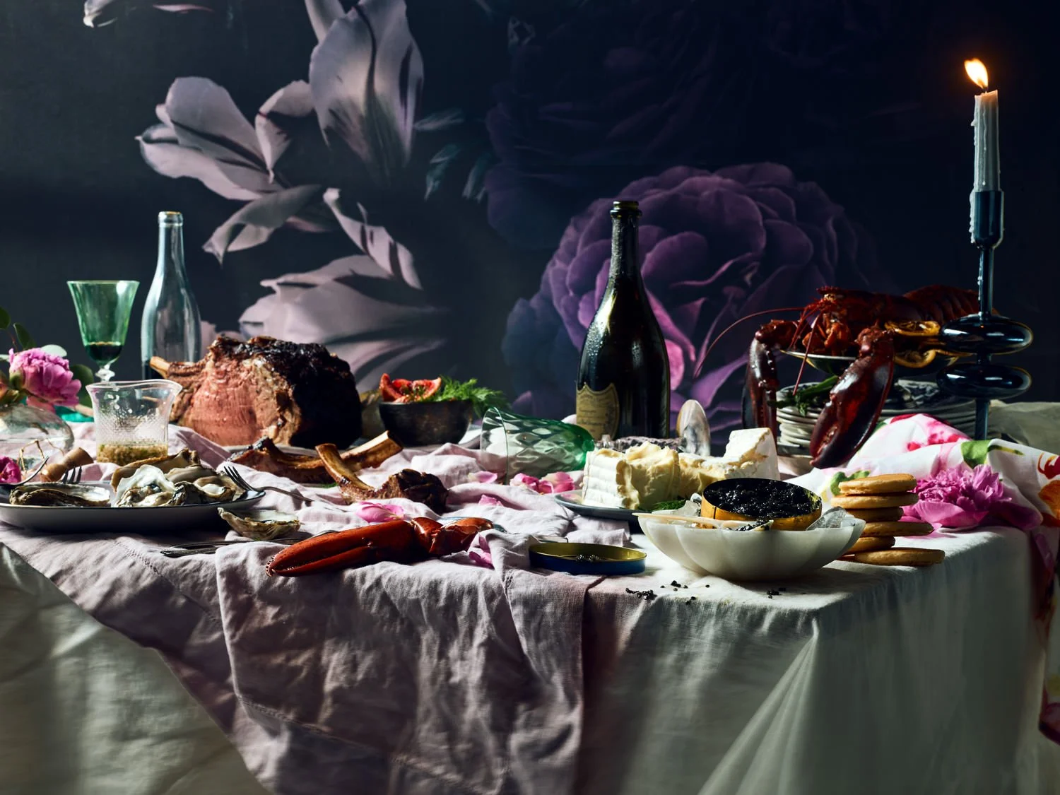 A table set for a feast with a large cut of meat, lobster, cheese, crackers, oysters, wine, and pink flowers, with a dark, floral-decorated background.