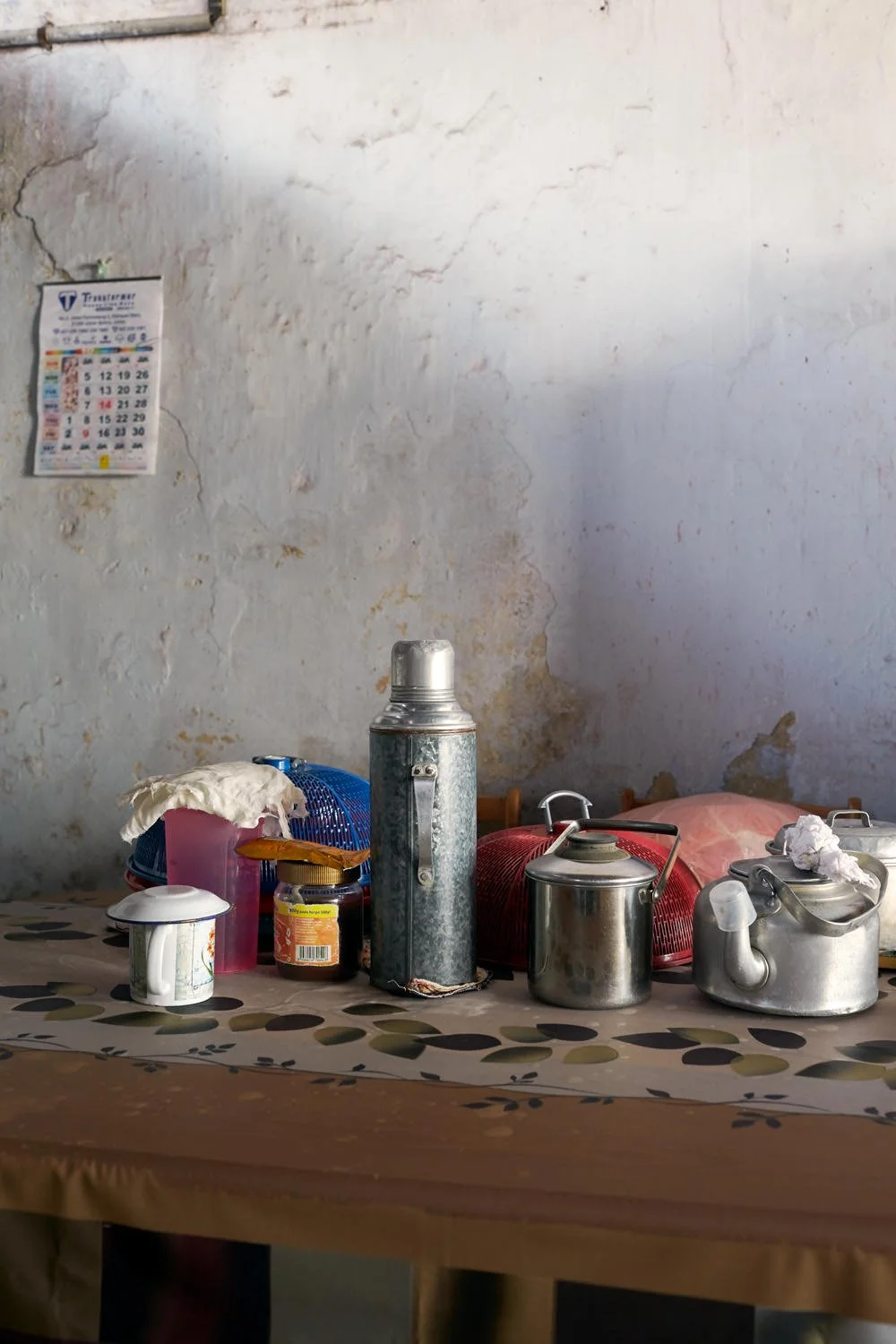 A cluttered kitchen table with various metal and plastic kitchen utensils, containers, and a thermos, against a weathered wall with peeling paint and a calendar.