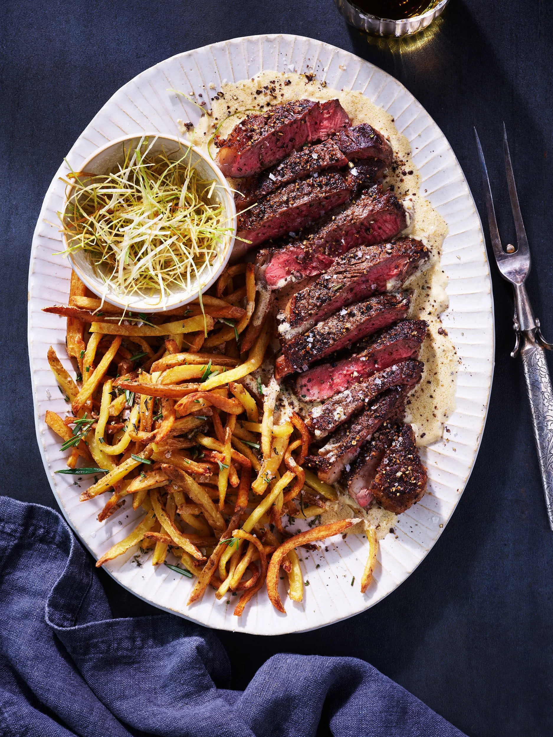 Sliced medium-rare steak with black pepper and salt, served with fried potato strips, a small bowl of microgreens, and a side of creamy sauce on a white oval plate.