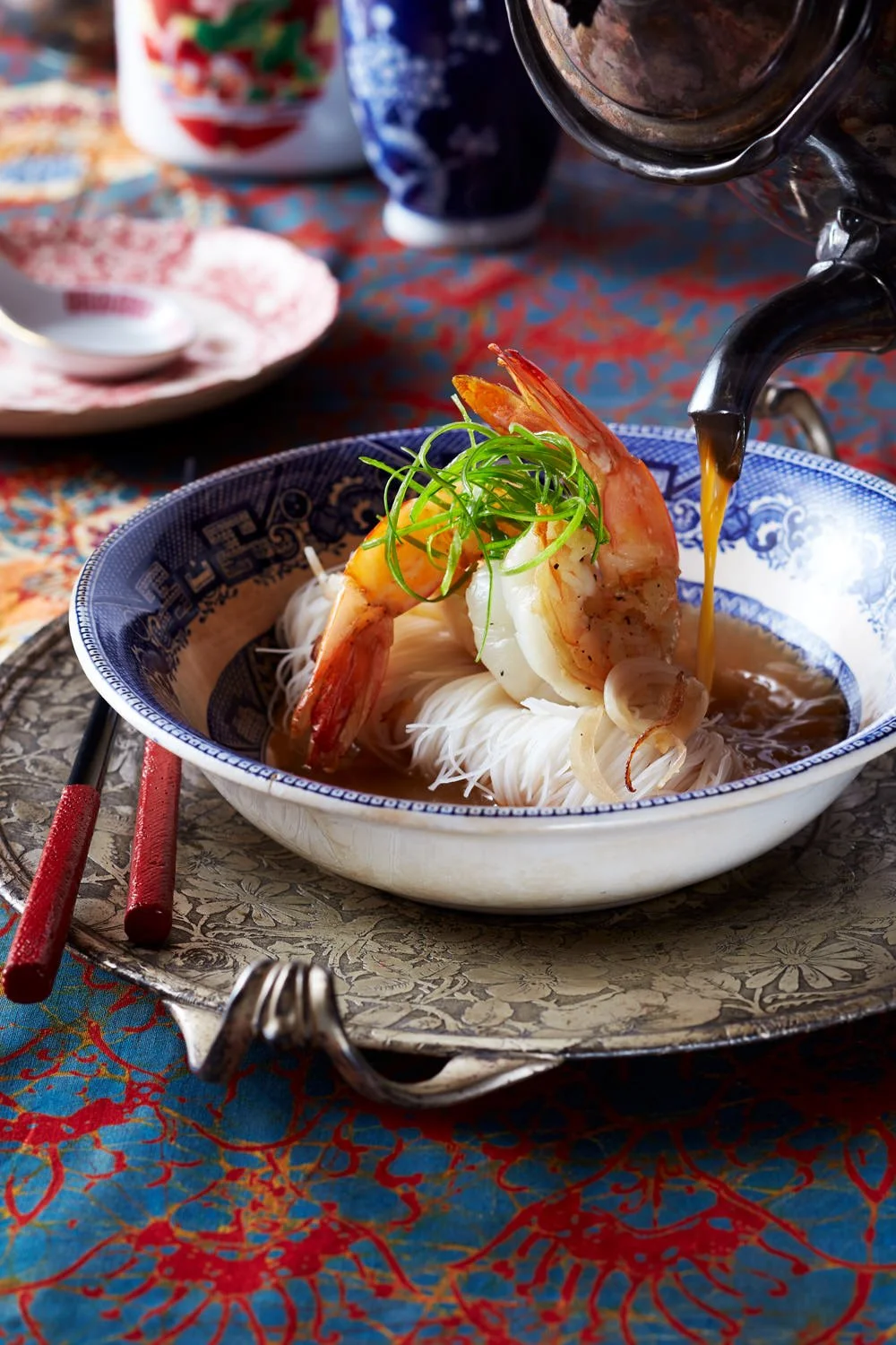 A bowl of Asian seafood soup with shrimp, noodles, and green onions, with broth and soy sauce being poured in, placed on a decorative tray with chopsticks and a fork, on a colorful patterned tablecloth.