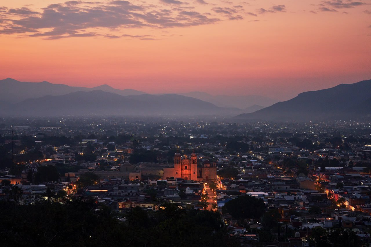 Cityscape at dusk with a sunset sky, mountains in the background, and illuminated streets and buildings, including a prominent historic church.