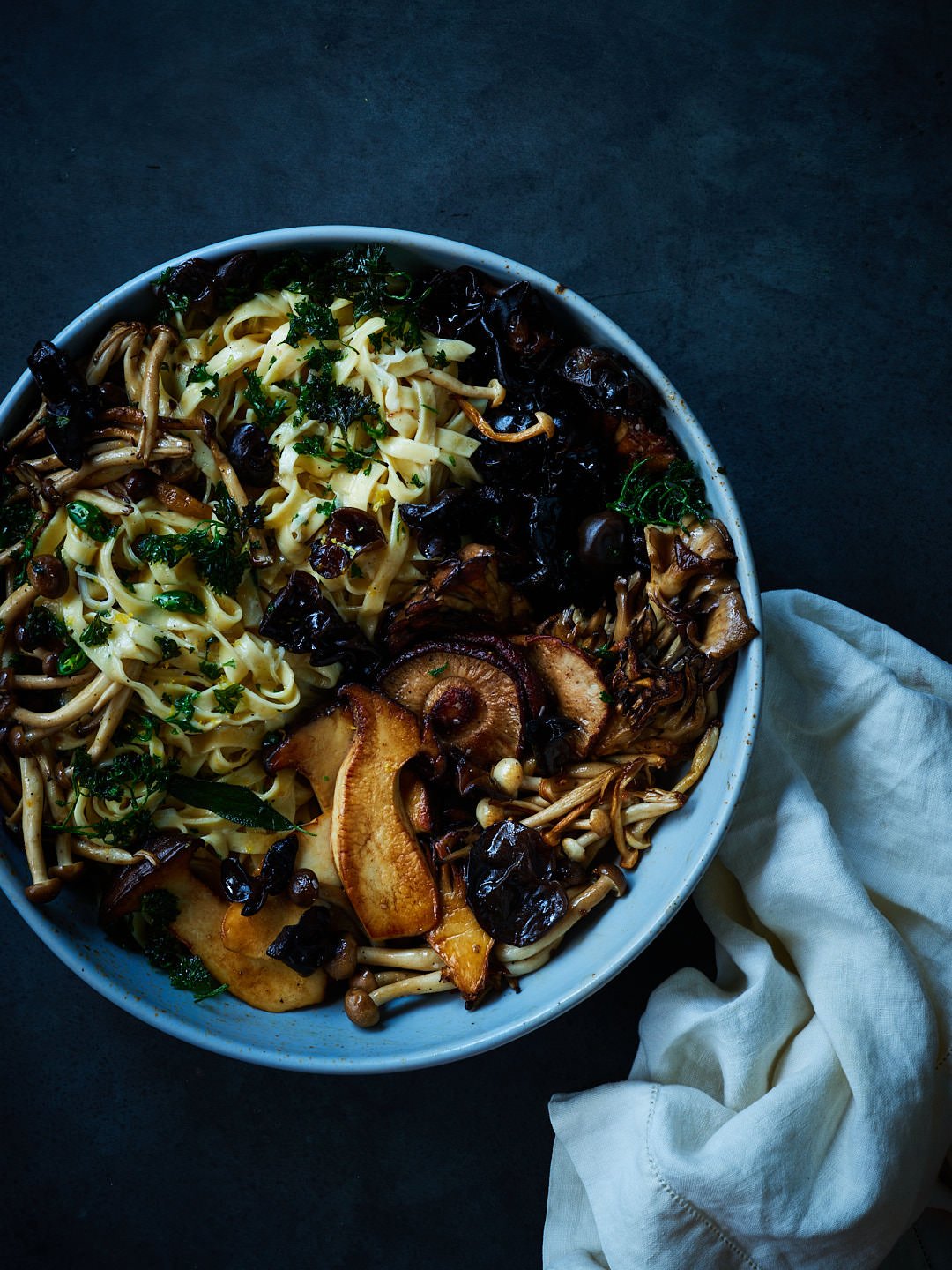 A bowl filled with various types of cooked mushrooms and pasta, garnished with chopped herbs, on a dark surface with a white cloth nearby.