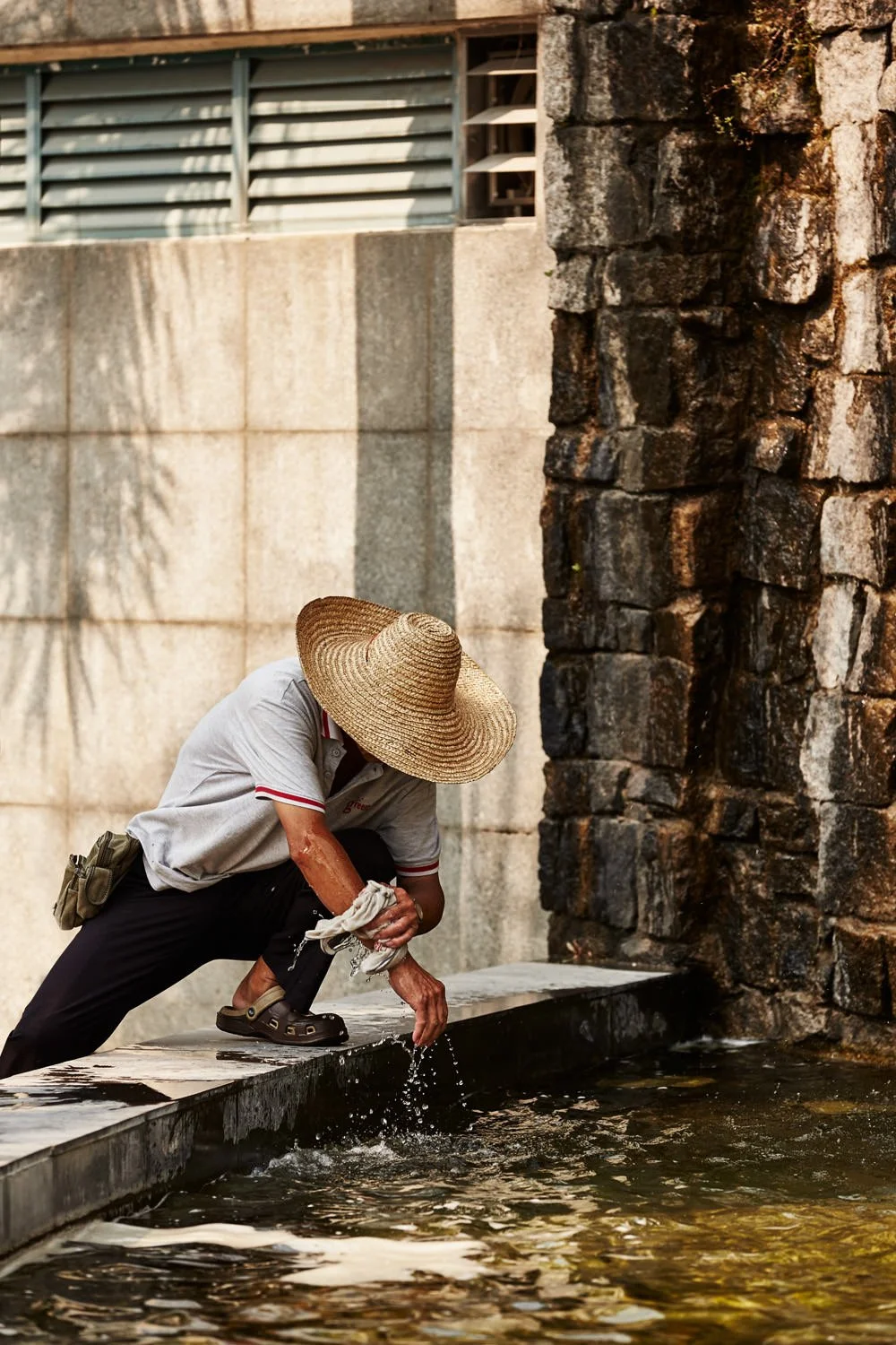 A person wearing a wide-brimmed straw hat, gray shirt, and black pants is crouching on a ledge by a water feature, wearing sandals, and holding a cloth while adjusting or washing their hands in the water.
