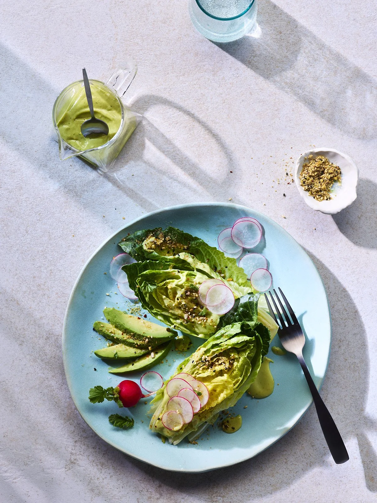 A plate of sliced avocado and lettuce with radish slices, a small bowl of crushed nuts, and a fork on a light-colored surface with sunlight shadows.