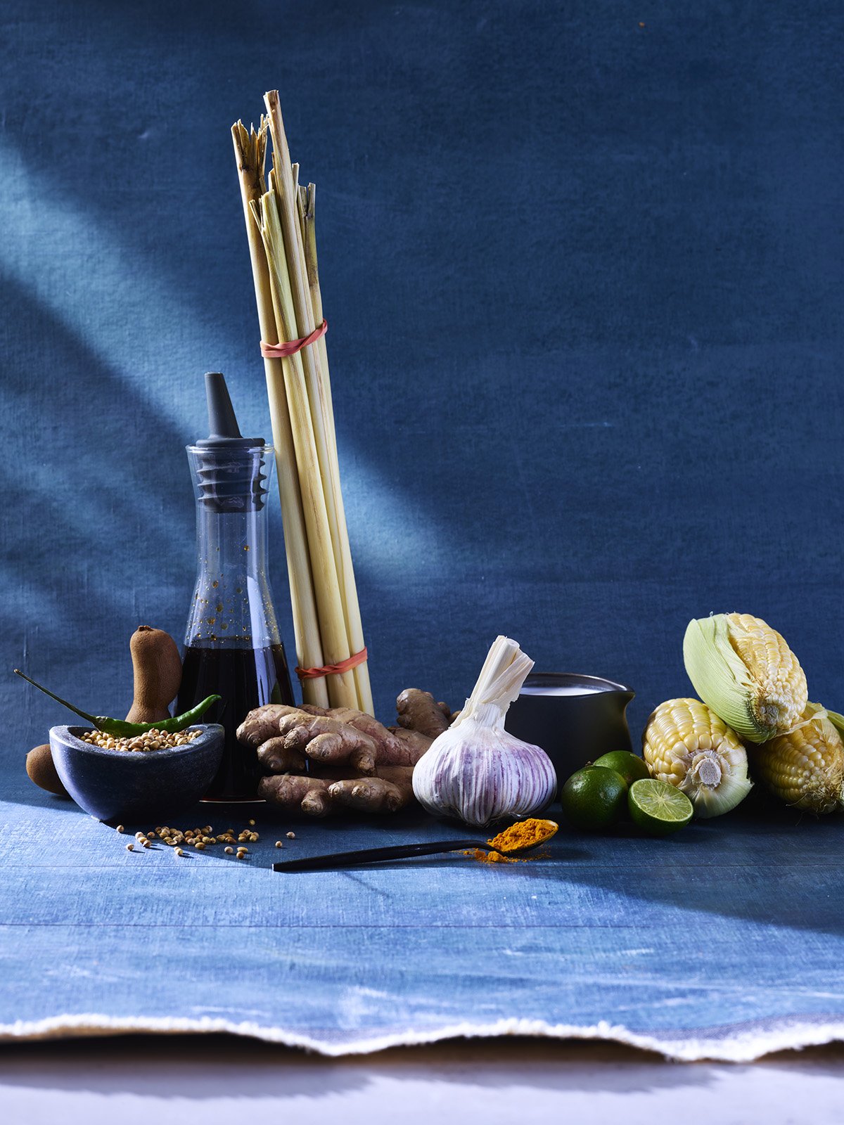 Assorted vegetables, herbs, and cooking ingredients on a blue surface with a dark blue background, including garlic, ginger, corn, lime, and spices.