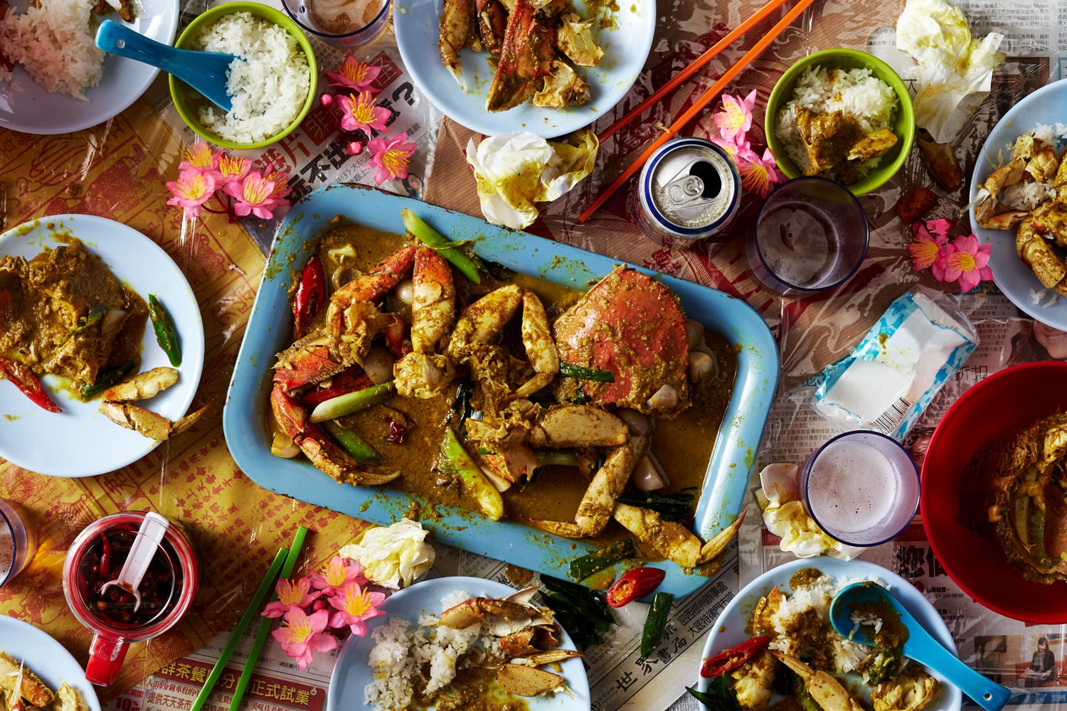 A table filled with various Asian dishes, including crab, rice, and curry, with cups, chopsticks, and decorative flowers.