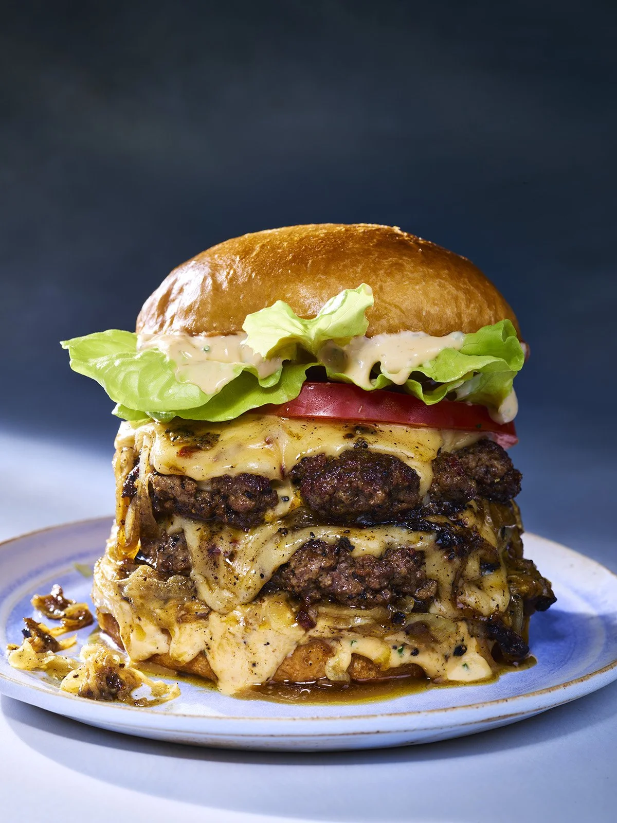 A towering burger with multiple layers of beef patties, melted cheese, lettuce, tomato, and sauce, served on a plate.