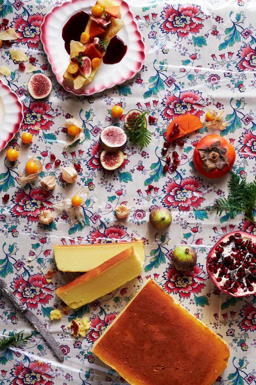 A floral-patterned tablecloth with assorted fruits, cheese, and desserts including figs, apples, persimmons, pomegranate, cheesecake, and a cake, some cut open and some whole, with scattered pomegranate seeds and other small fruits.