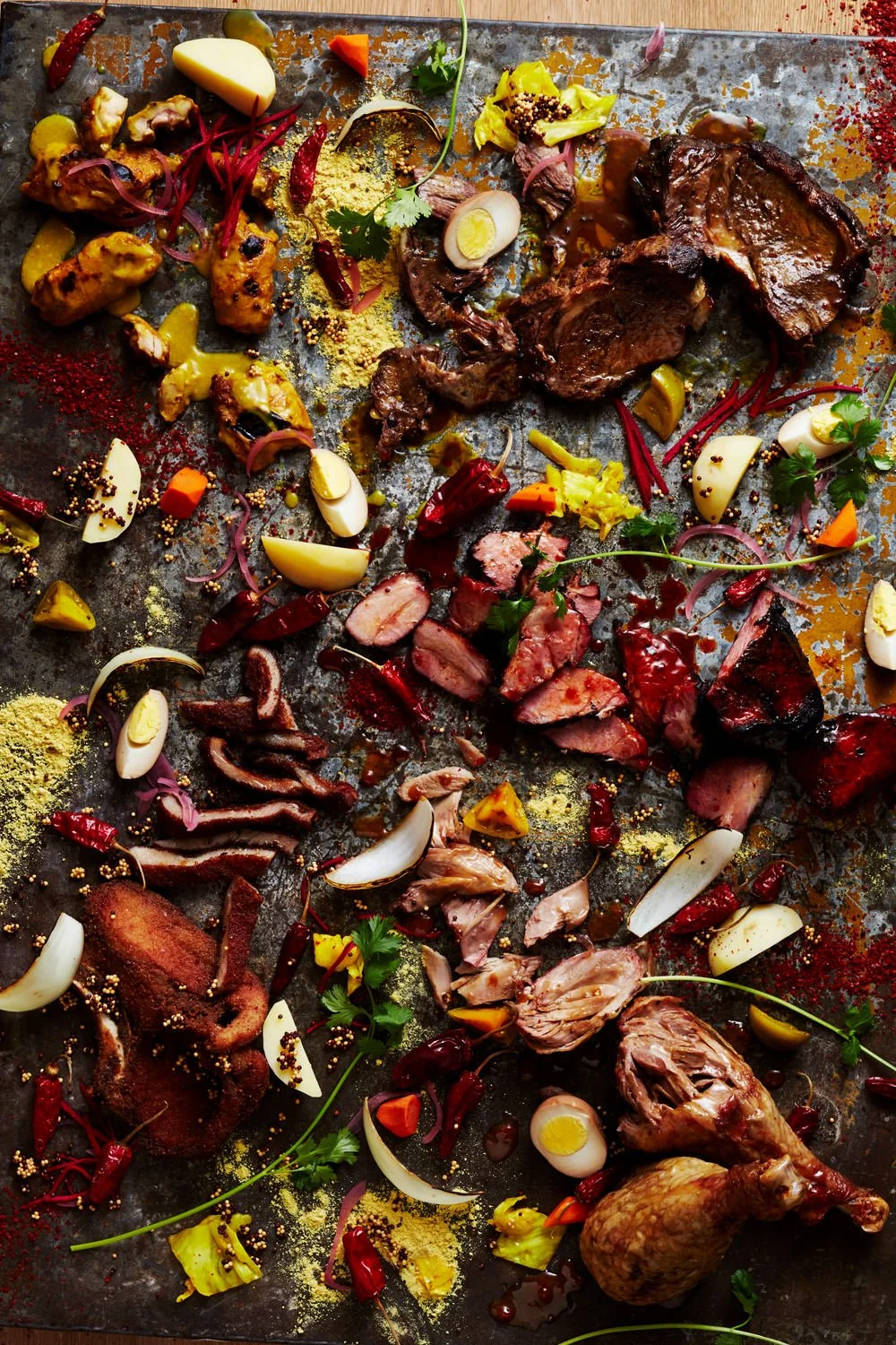 Assorted grilled and roasted meats along with chopped vegetables, hard-boiled eggs, herbs, and spices on a metal surface.
