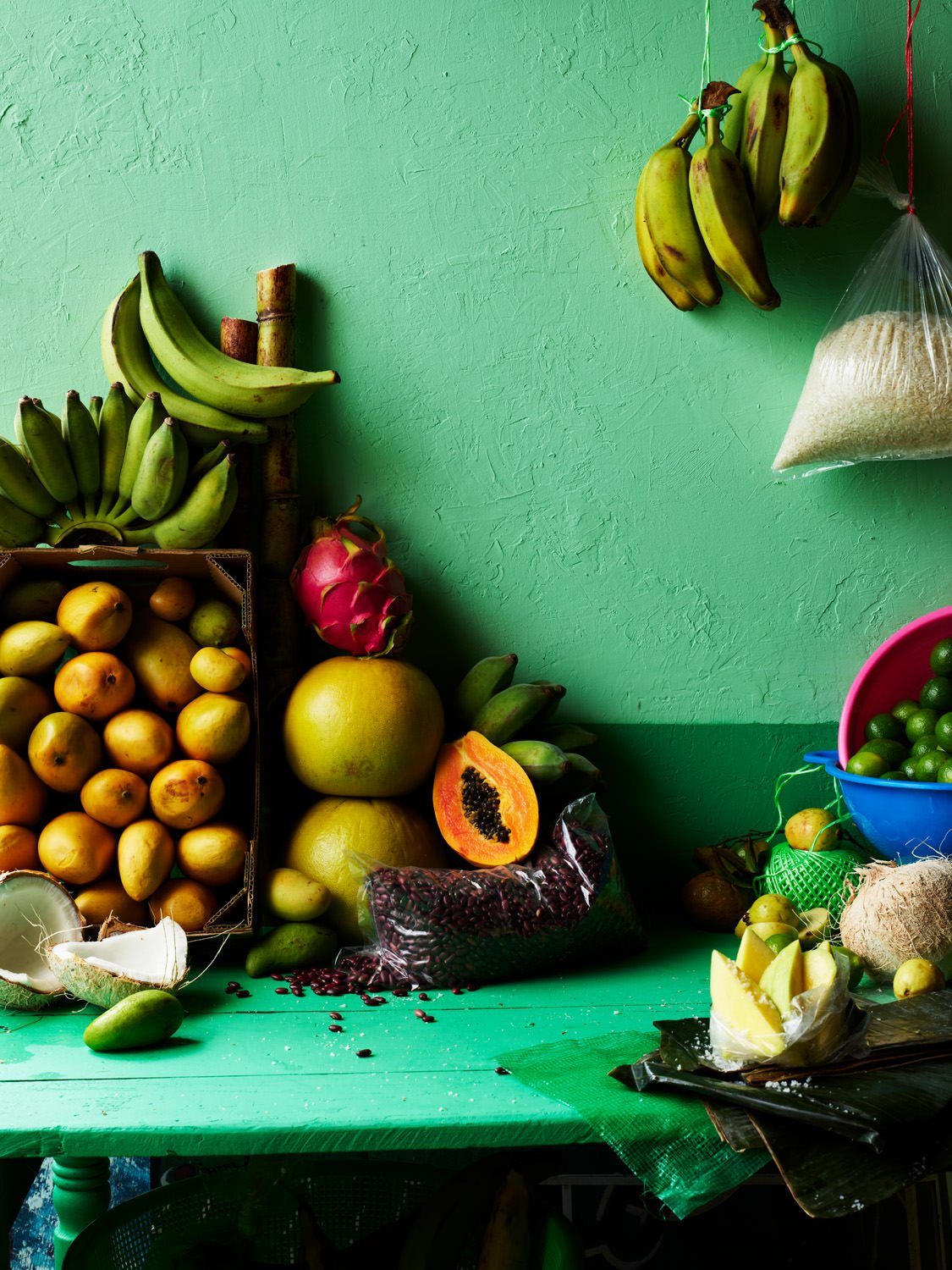 A variety of fresh tropical fruits on a green table and wall, including bananas, papaya, dragon fruit, lime, coconut, and a bowl of small green limes.