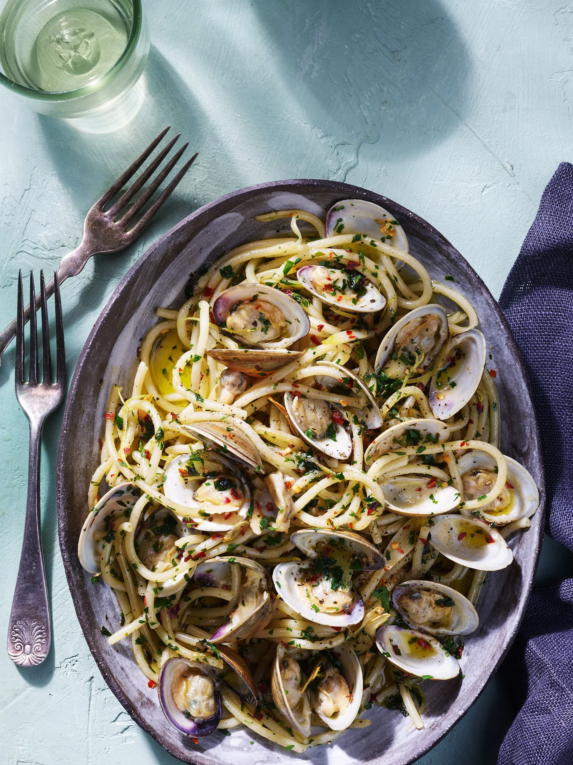 A plate of spaghetti with clams and herbs on a rustic dish, accompanied by two forks, a glass of water, and a dark napkin on a aqua textured table.