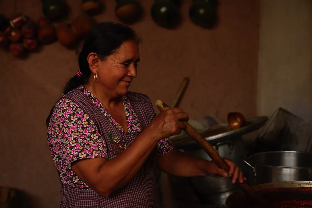A woman smiling as she stirs a pot with a wooden spoon in a rustic kitchen setting.