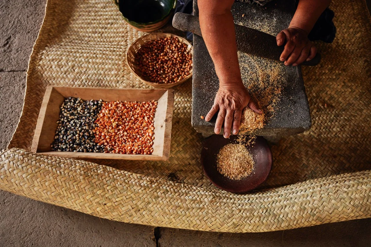 A person grinding corn using a traditional stone mill on a woven mat, with bowls of dried corn seeds and ground corn nearby.