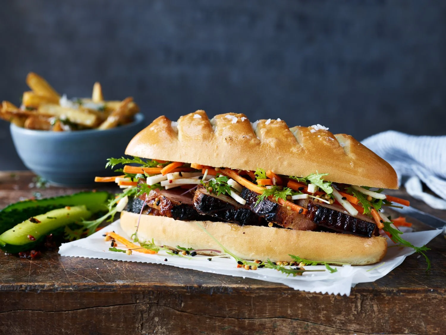 Sandwich with smoked meat, coleslaw, and pickles on a bun, served on a wooden surface with a side of fries in a bowl.