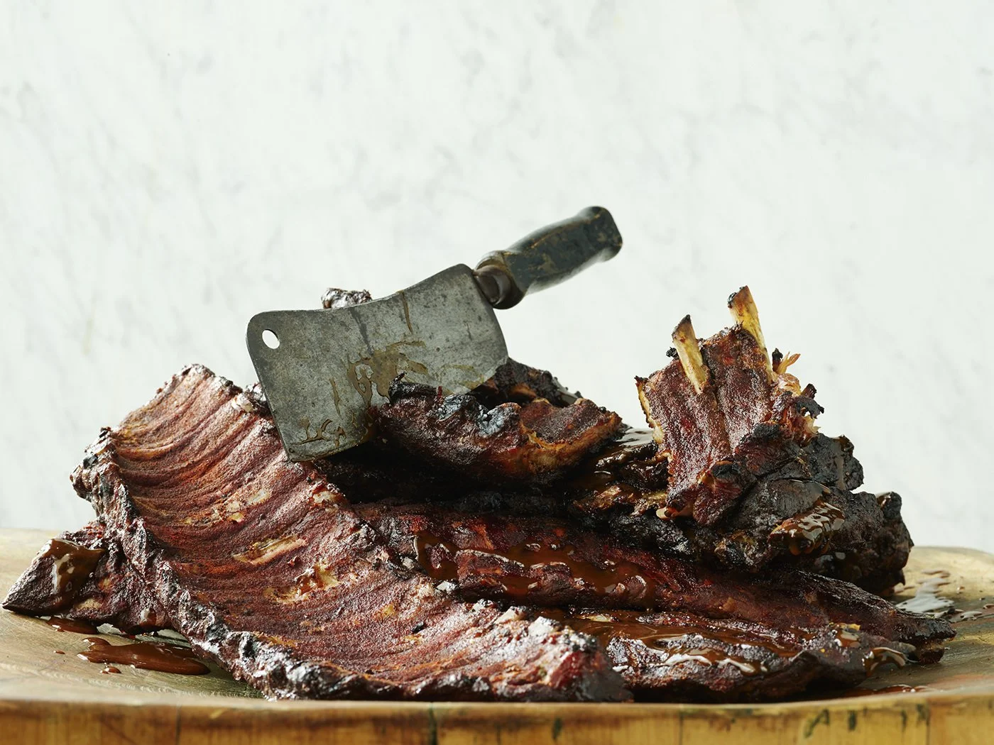 A large pile of cooked barbecue ribs on a wooden surface with a meat cleaver placed on top.