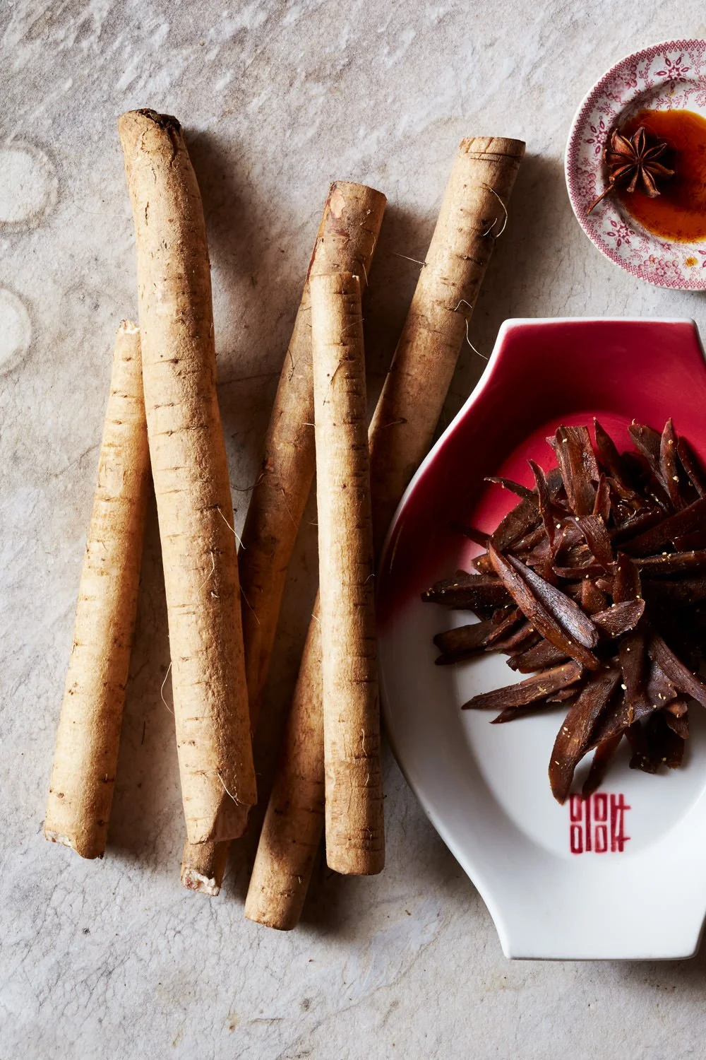 Ginseng roots, a small dish of dark extract or tea with star anise, and a white dish with sliced dried jujube or another dried fruit on a textured surface.