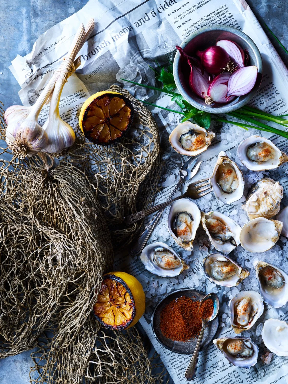 A rustic seafood scene featuring fresh oysters on a bed of ice, grilled lemon halves, garlic bulbs, red onion, chili powder, and a crumpled newspaper.