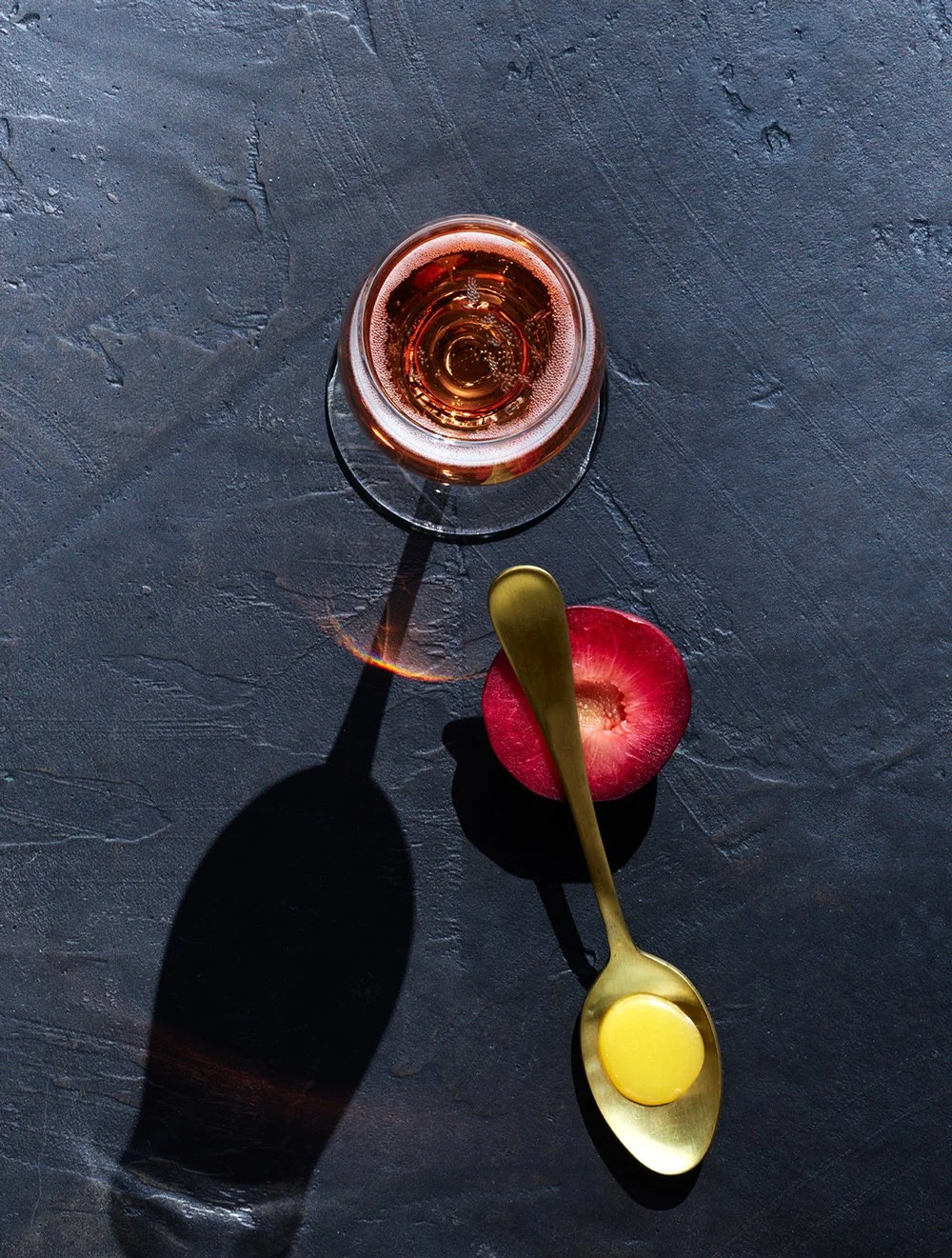 Top-down view of a glass of rosé wine, a halved peach with a gold spoon holding honey or egg yolk on it, all on a dark textured surface.