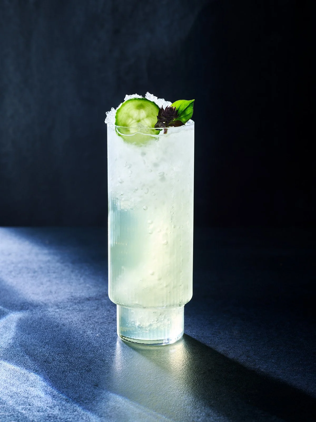 A tall glass of a clear cocktail garnished with a cucumber slice, basil leaves, and some salt on the rim, set against a dark background.