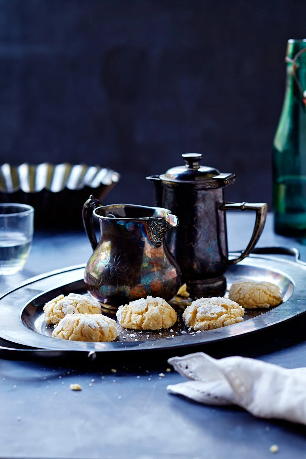 A vintage tray with five cookies and a coffee pot, a creamer, a glass of water, and a green bottle on a blue table with a dark background.