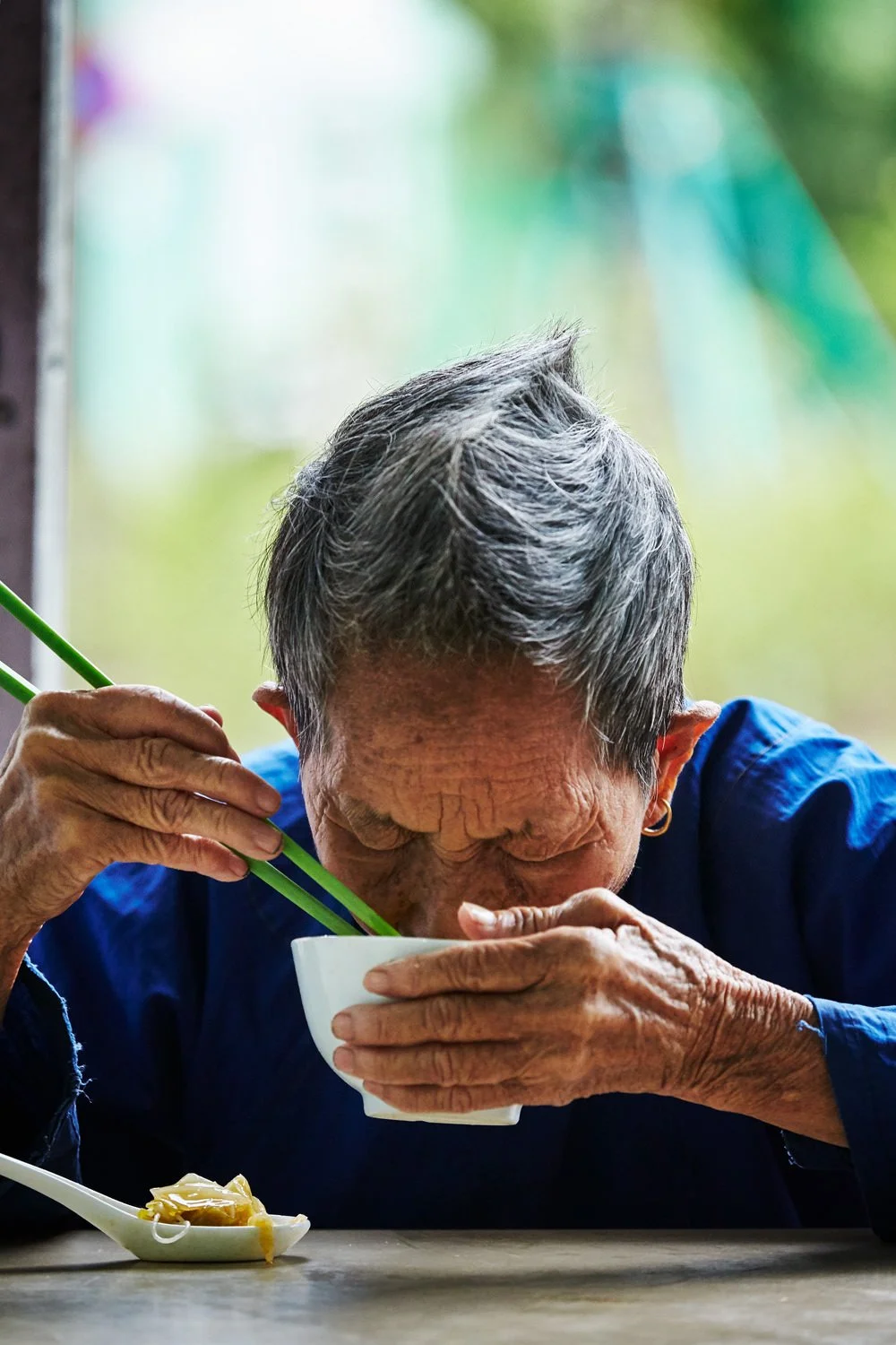 An elderly woman with gray hair and earrings drinking from a white bowl using green chopsticks, sitting at a table with a spoonful of noodles nearby.