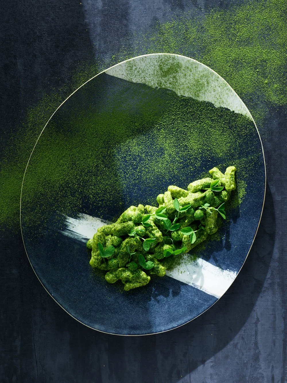 Creative food presentation with green elements, including a textured green garnish or edible item adorned with small green leaves on a black, round plate with green powder or seasoning scattered for decorative effect.