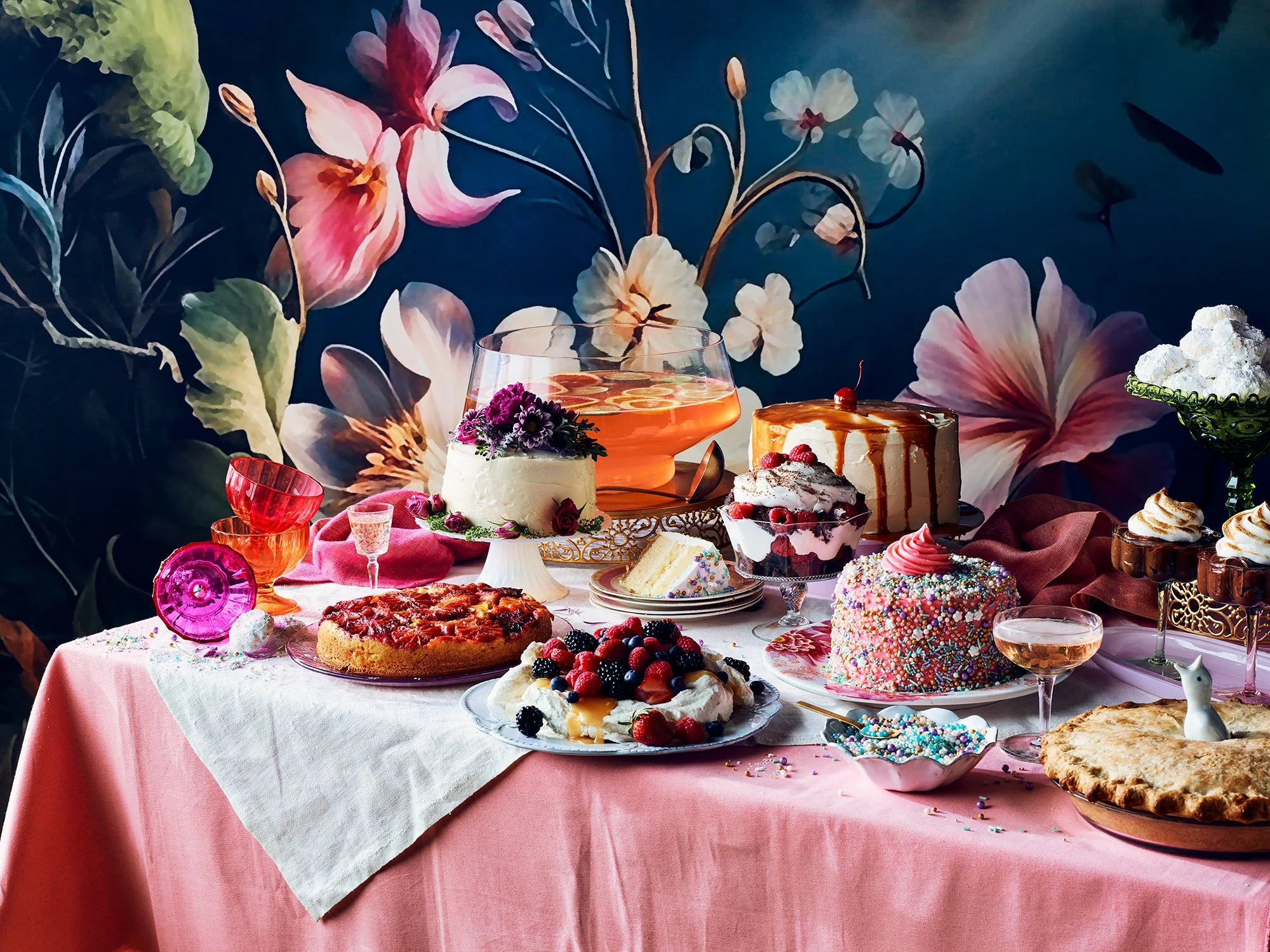 A variety of desserts and drinks on a pink tablecloth, including cakes, pies, a fruit tart, and colorful glassware, with a floral mural background.