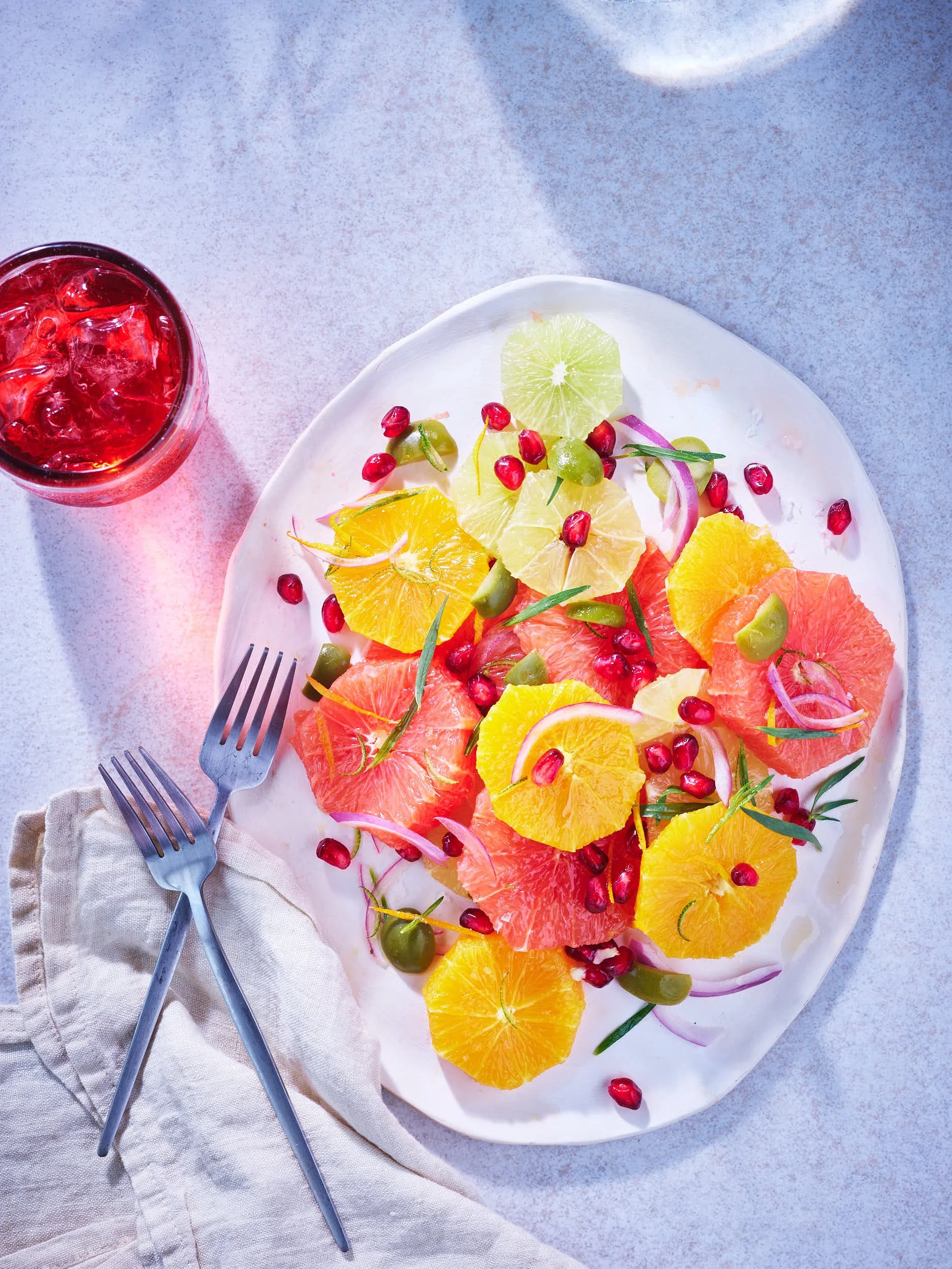 A colorful grapefruit and orange fruit salad on a white platter with pomegranate seeds, onion slices, and green olives, accompanied by a glass of red drink with ice and a pair of stainless steel forks on a beige napkin.