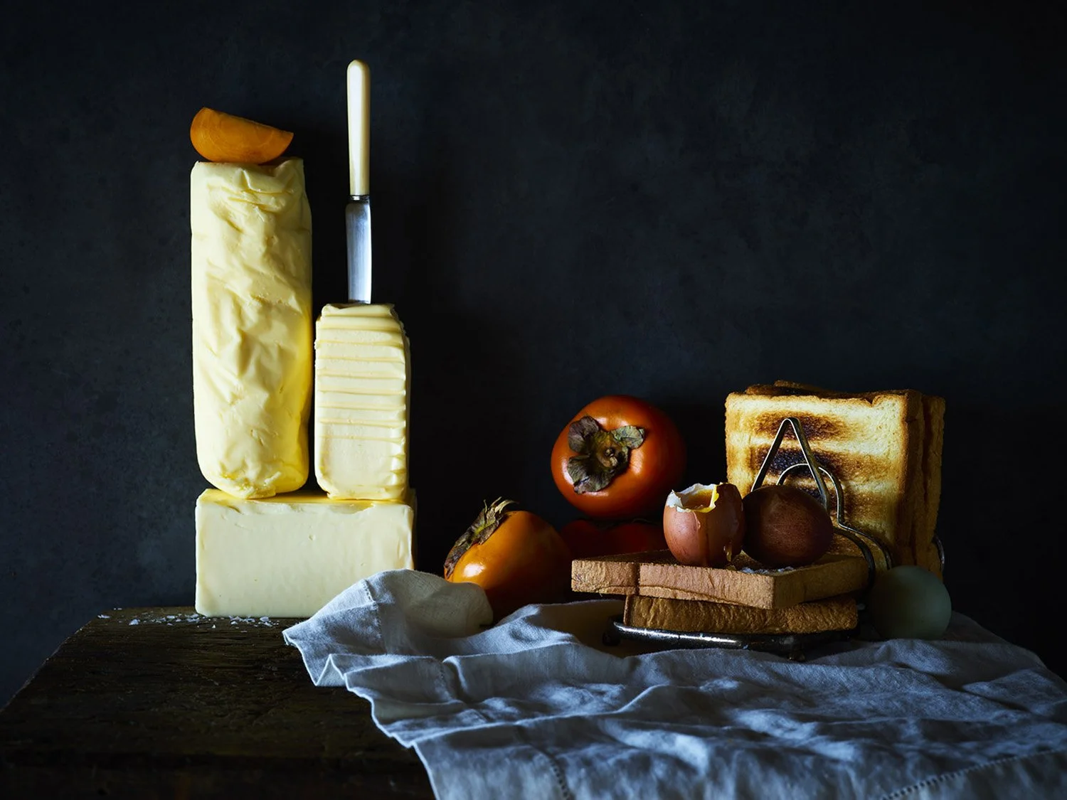 Various cheeses, persimmons, bread, and eggs arranged on a dark surface with a cloth, with a knife inserted into a block of cheese.