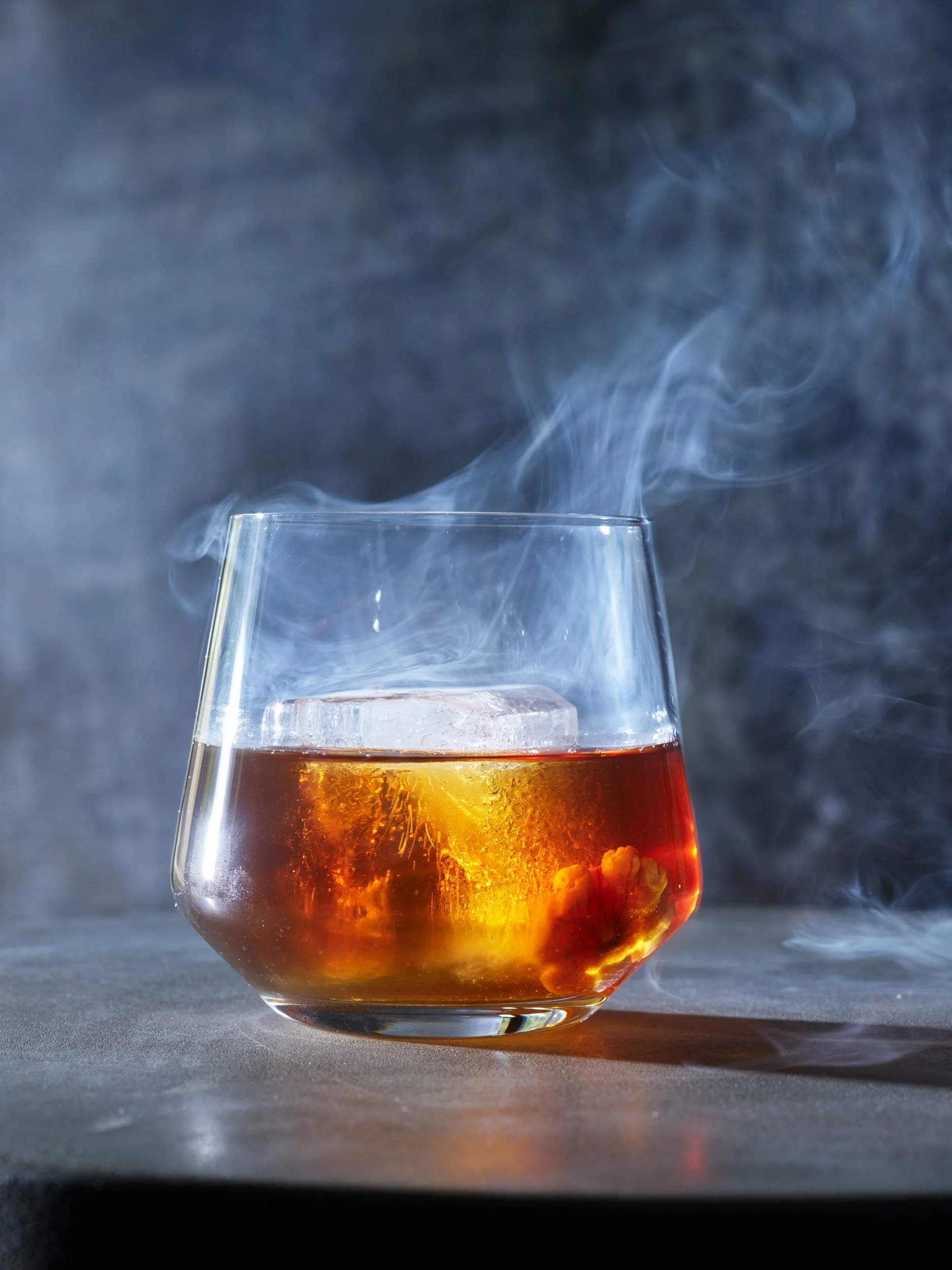 A glass with amber-colored alcohol and ice, with smoke swirling around it on a dark surface.
