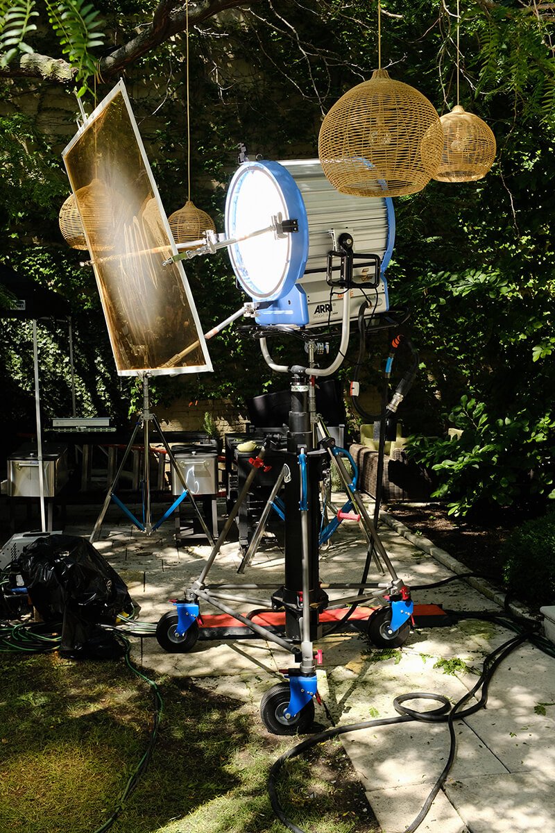 Professional film or video lighting equipment set up outdoors, including a large light with a reflector, a circular light, and wicker pendant lamps hanging from trees.