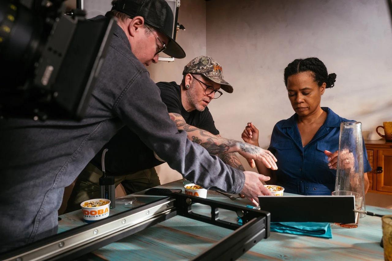 People filming a scene with food, including a woman in a blue shirt, a man with glasses and tattoos, and another person wearing a cap, around a table with bowls of food.