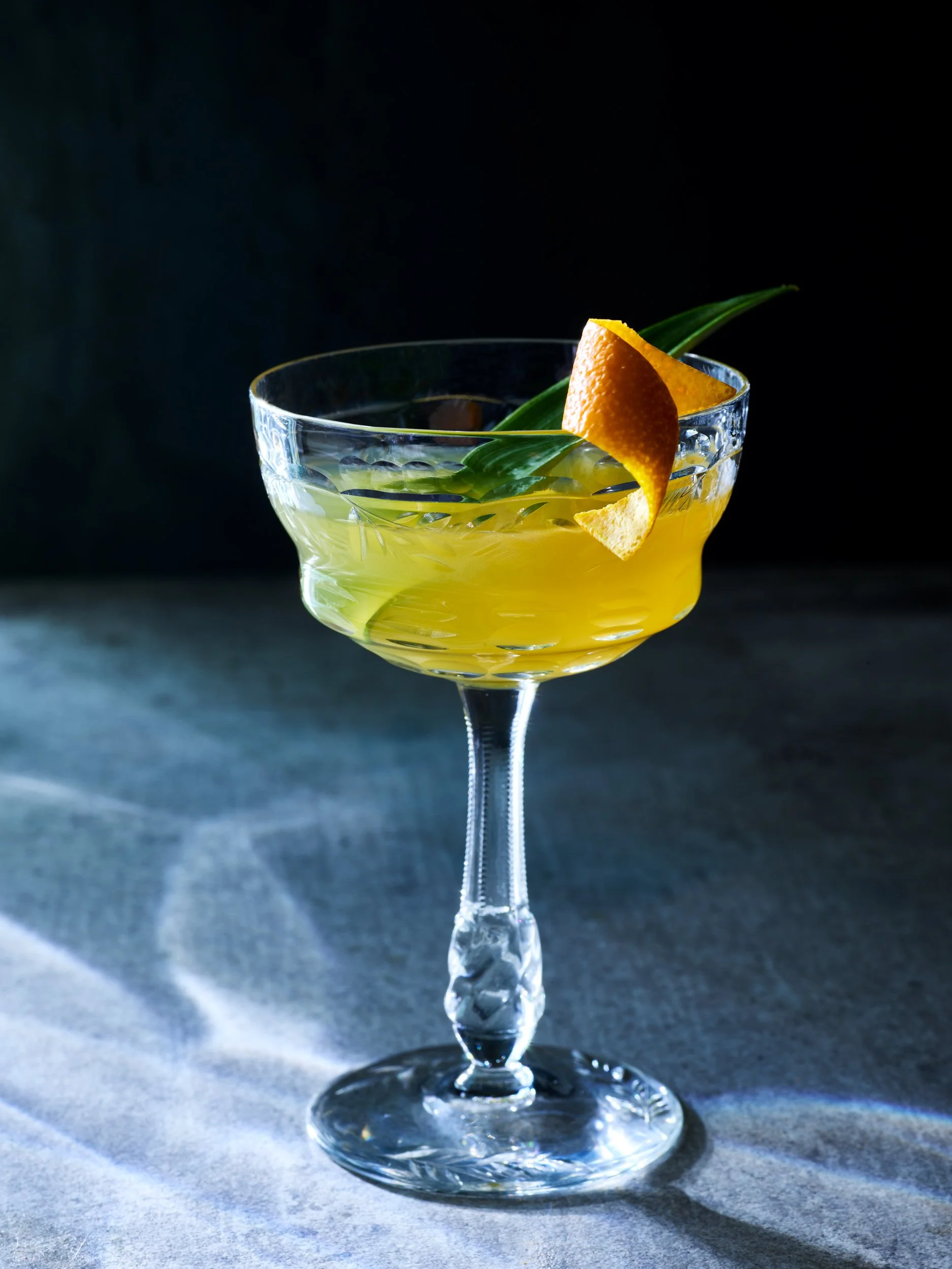 A glass cocktail with yellow liquid, garnished with an orange twist and green leaves, on a dark surface.