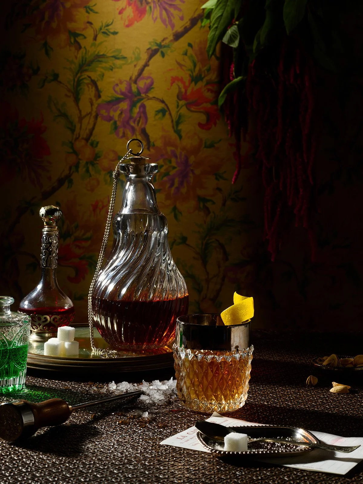A bar setting with a crystal decanter of red beverage, a glass filled with the same drink garnished with a lemon peel, and various bottles and drinks on a dark textured table. The background features a floral wallpaper with yellow, purple, and green 