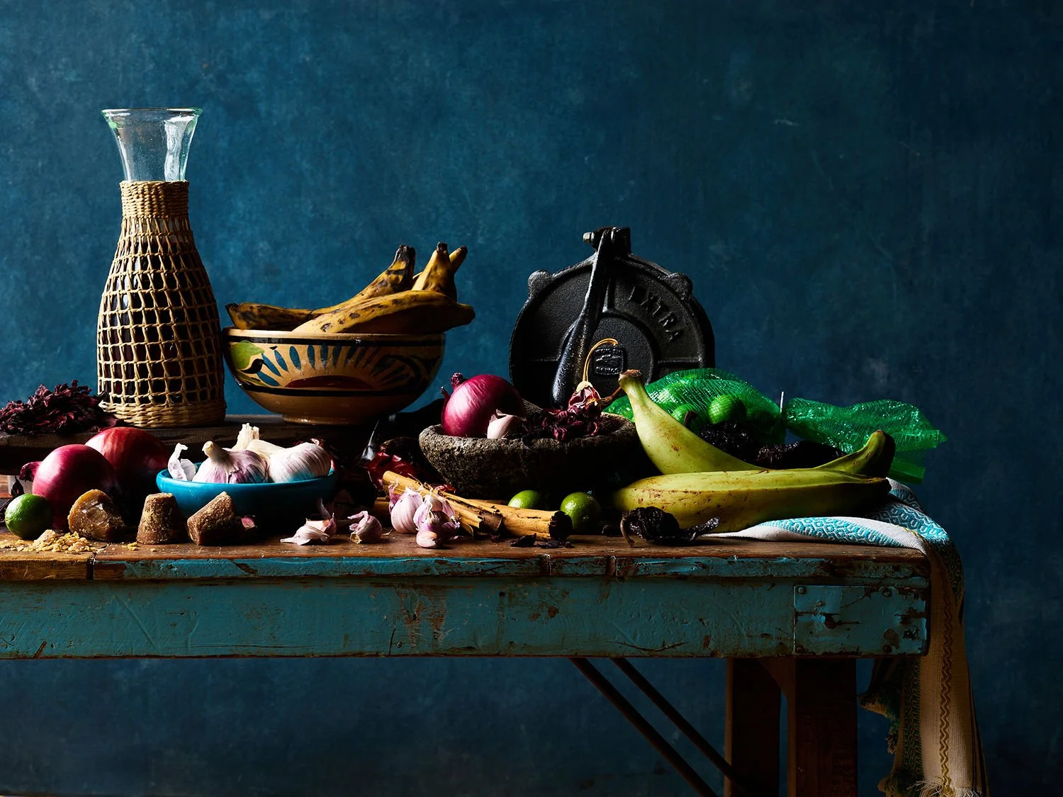 A rustic wooden table with various vegetables, fruits, garlic, and kitchenware against a dark blue background.